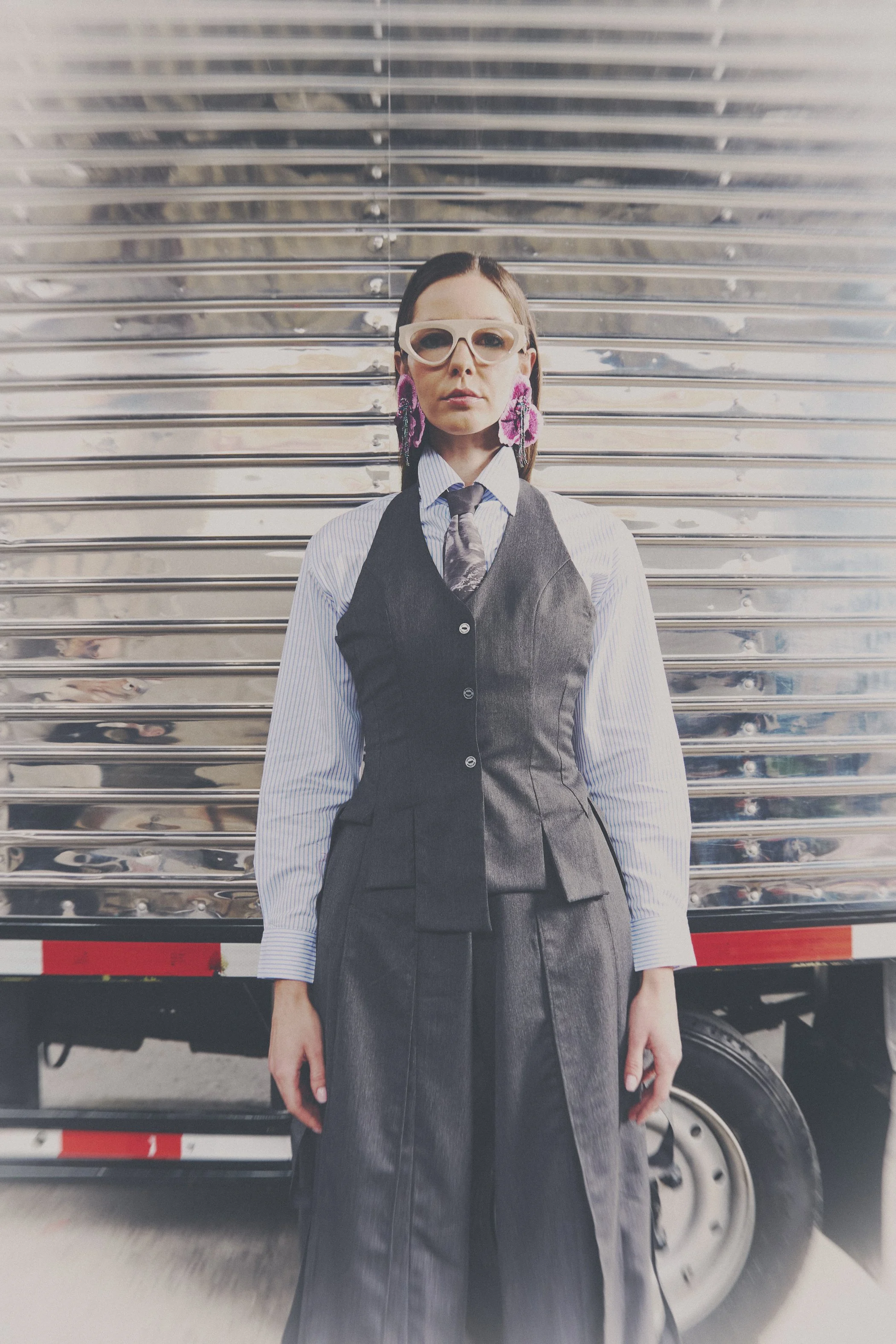 Mujer con gafas grandes, camisa azul con rayas, chaleco negro, pantalones de mezclilla, y pendientes rosas, de pie frente a un camión de metal.