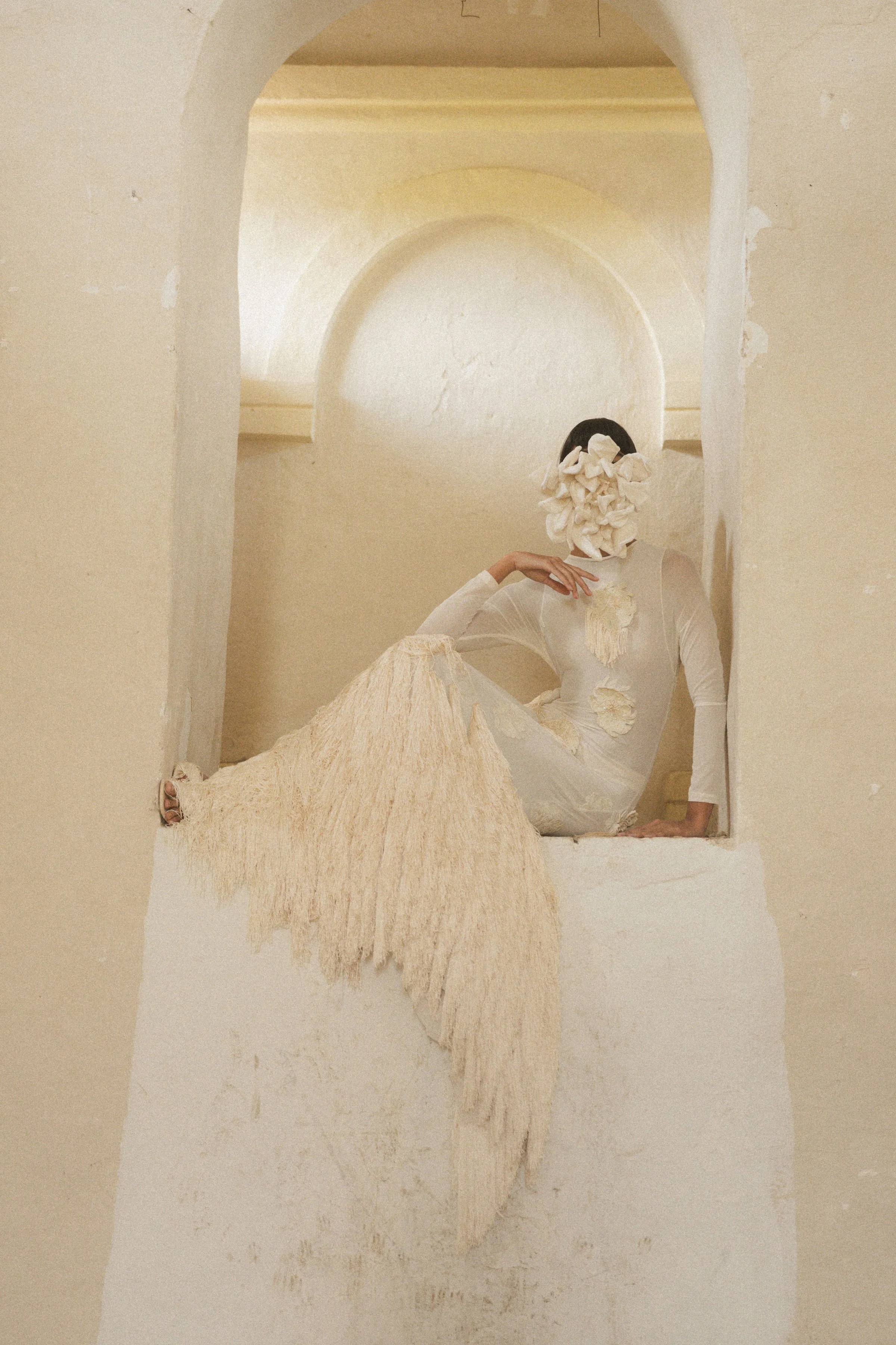 Mujer con rostro cubierto por telas blancas, vestida con un vestido largo y una capa de flecos en tono claro, sentada en una ventana en una pared beige.