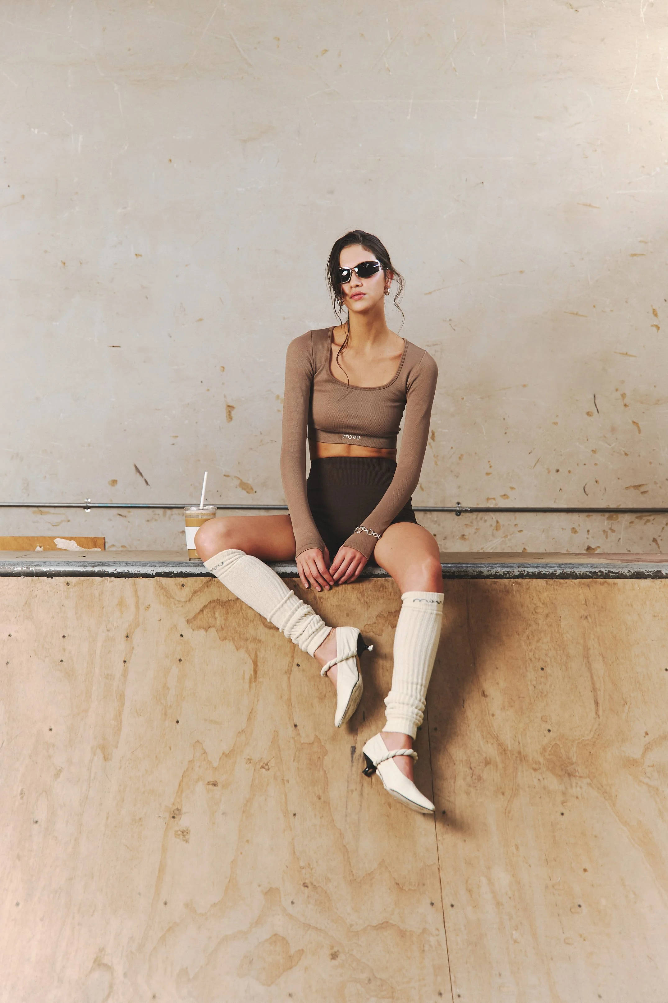 Joven mujer con gafas oscuras, camiseta ajustada de manga larga y falda negra, sentado sobre un borde de madera, con bebidas en la mano. Usa calcetas largas blancas y tacones blancos con tirantes.