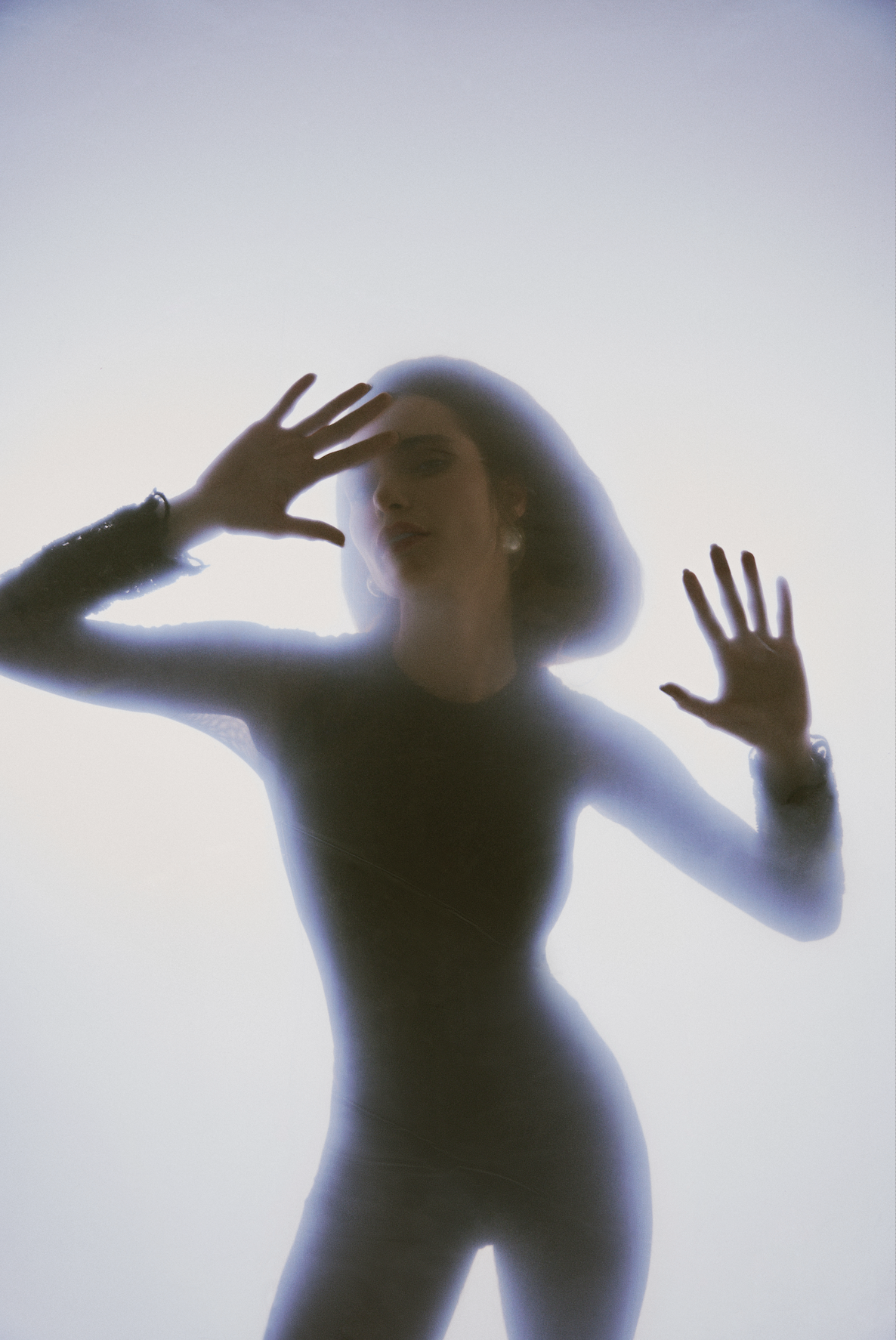 Mujer con sombrero y ropa ajustada, vista detrás de un cristal transparente con neblina y luz suave, manos en la superficie