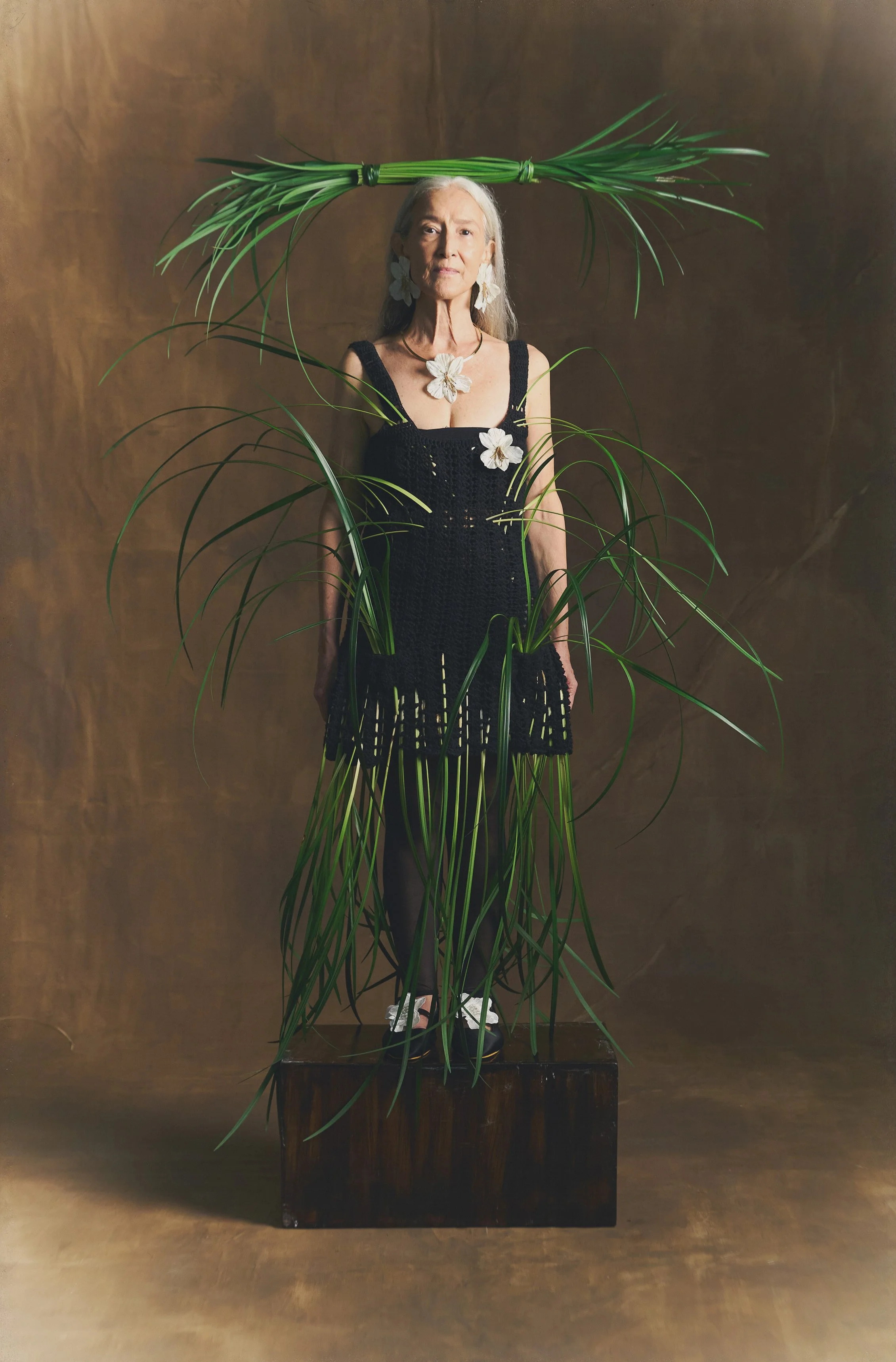 Mujer mayor vestida con un vestido negro y accesorios de flores blancas, rodeada y adornada con plantas verdes, posando sobre una plataforma de madera en un fondo marrón.