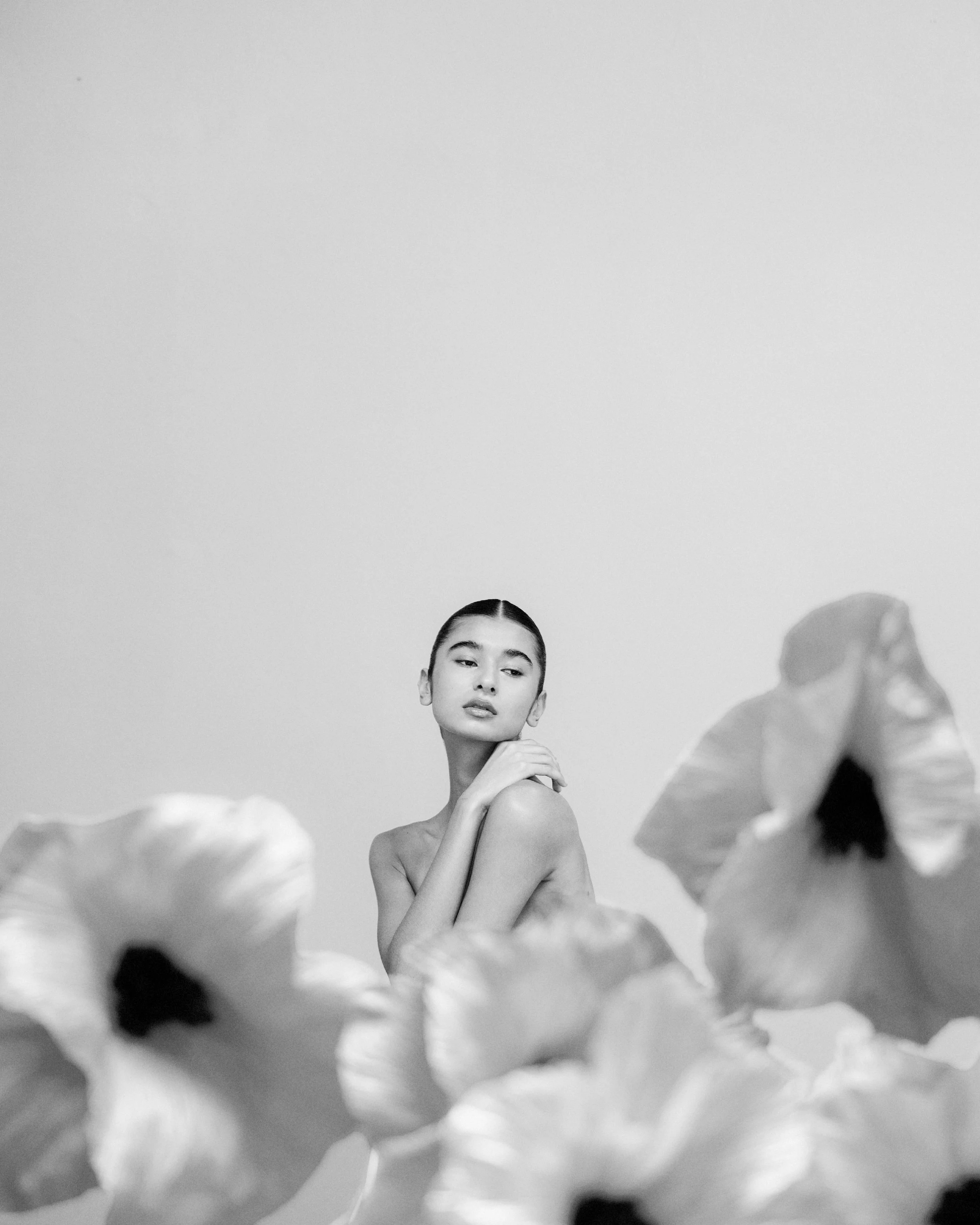 Retrato en blanco y negro de una mujer joven con cabello liso y peinado hacia atrás, rodeada de flores grandes y ligeras, con expresión serena y mirada hacia el lado.