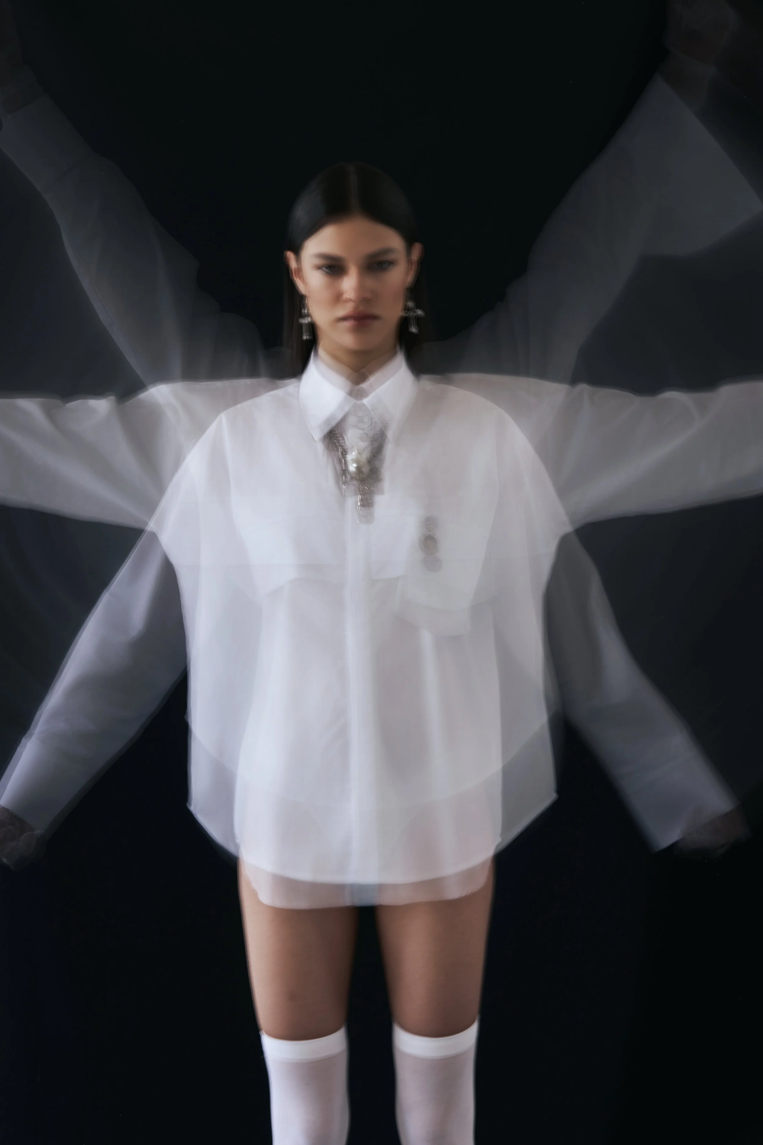 Mujer con vestimenta blanca y medias blancas en postura con brazos extendidos hacia arriba, movimiento borroso que crea un efecto de duplicación