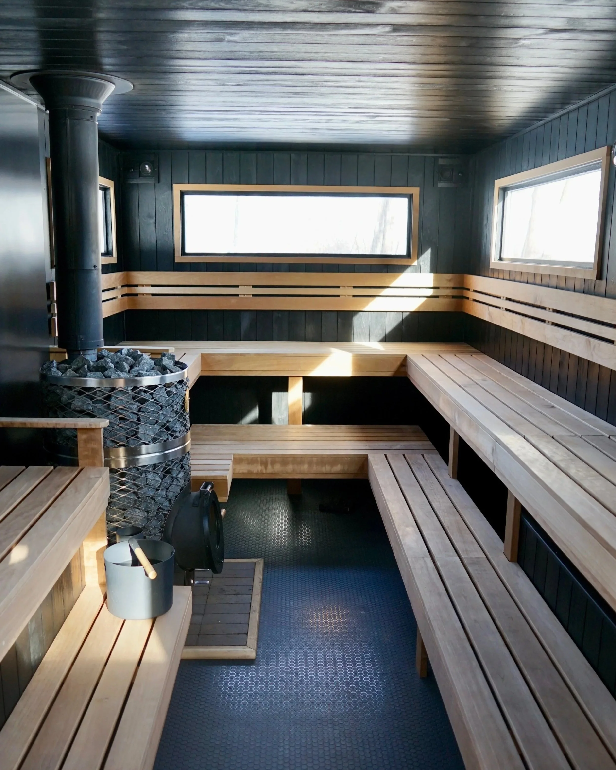Interior of a sauna with wooden benches, a stove with stones, and windows letting in natural light.
