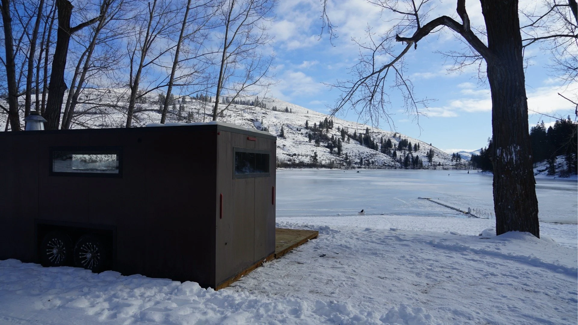 Snow-covered lake with a wooden shed and bare trees in the foreground, snow-covered mountains in the background, and a partly cloudy sky.