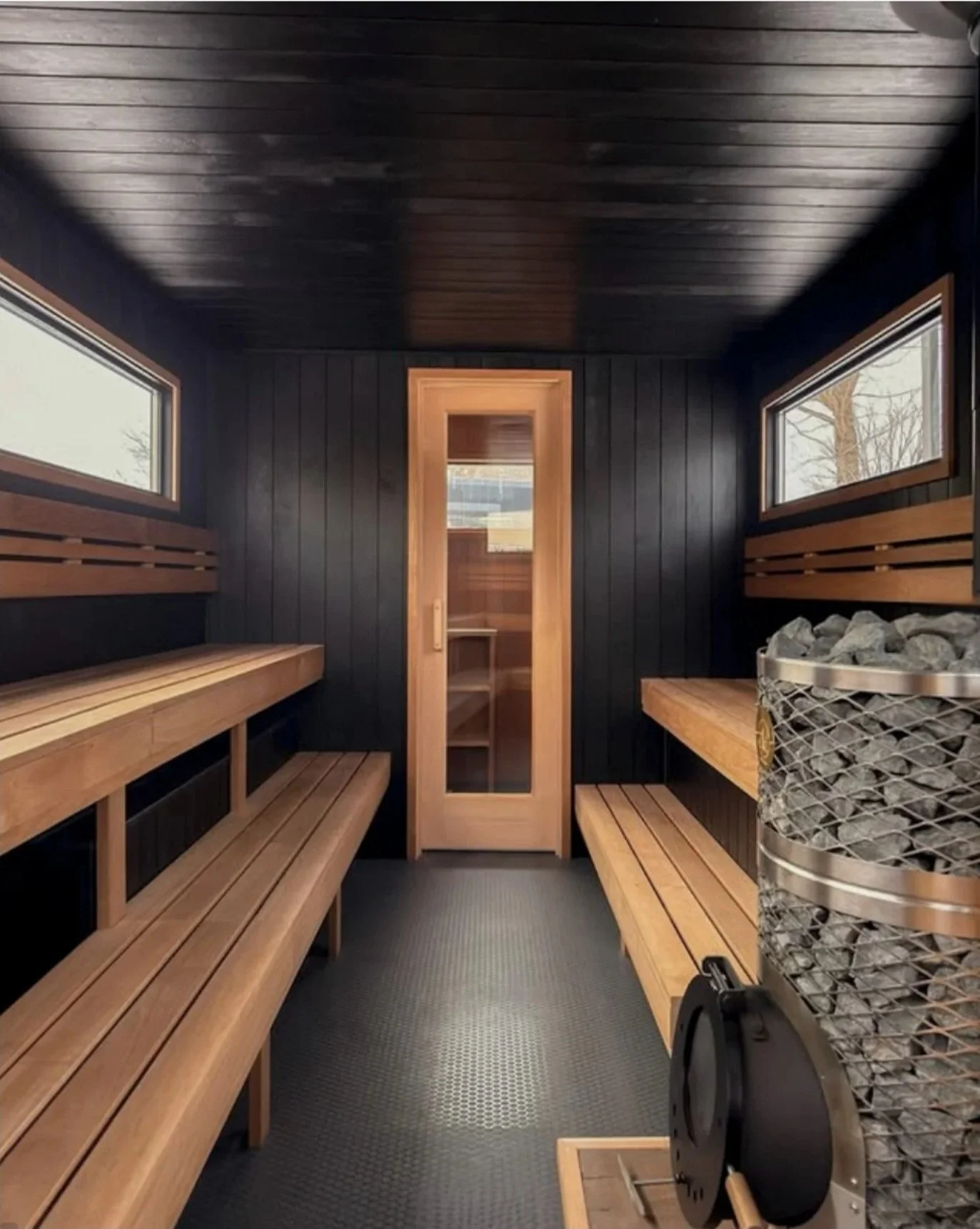 A small, modern sauna room with wood paneling. It has two long wooden benches on each side, small windows near the ceiling, and a door at the far end. There is a sauna heater with rocks on the right side.