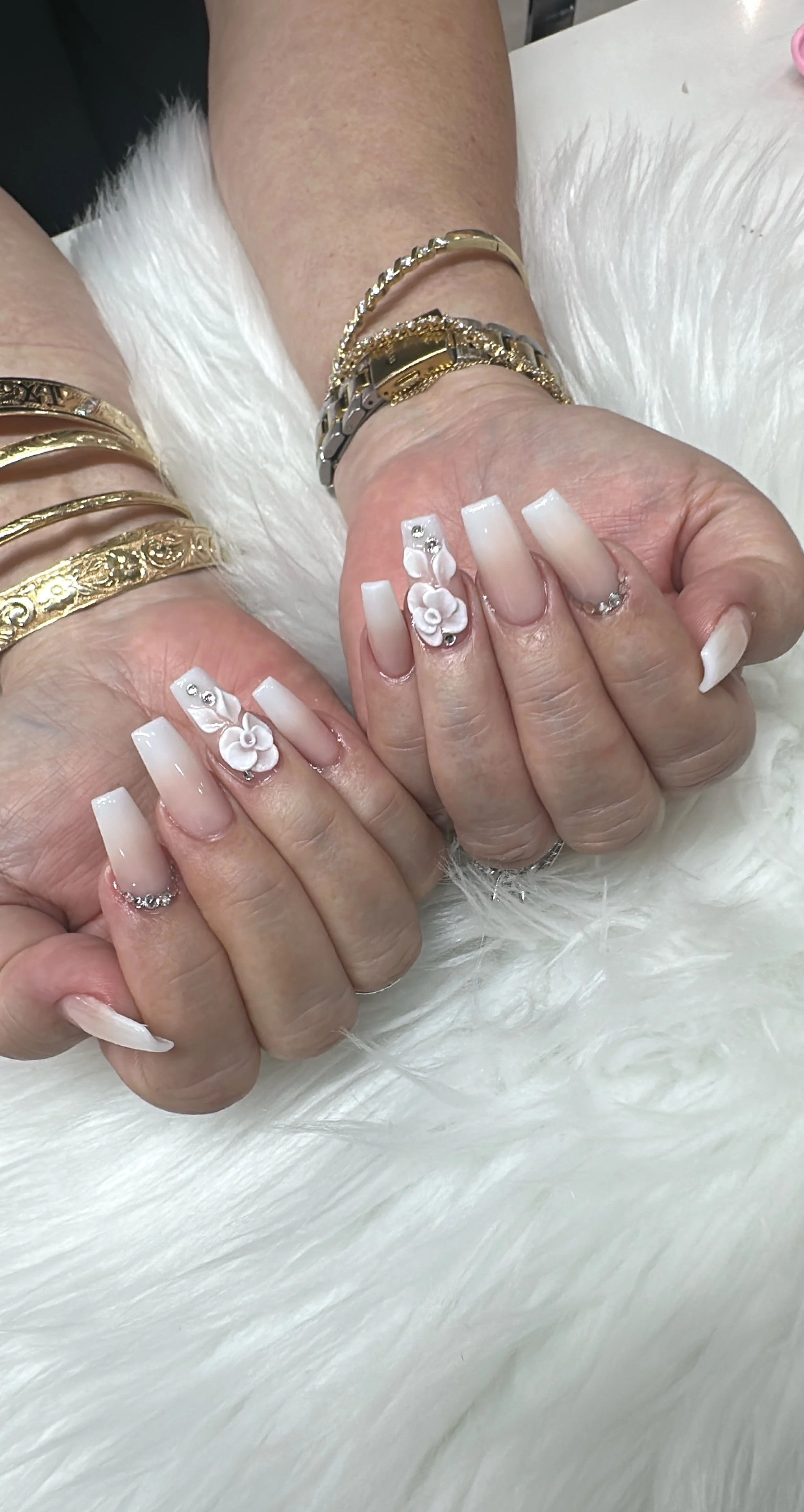 Close-up of a woman's hands with manicured nails featuring white polish, 3D floral embellishments, and rhinestones, resting on a white fluffy surface. She wears multiple gold and silver bracelets on both wrists.