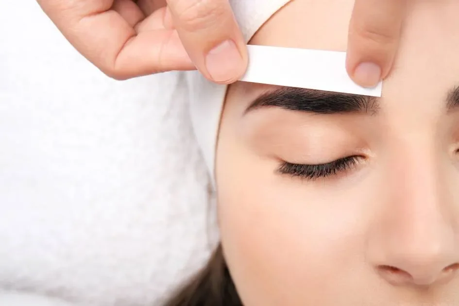 A person with dark hair and well-groomed eyebrows having a strip of waxing strip applied to their eyebrows for hair removal.
