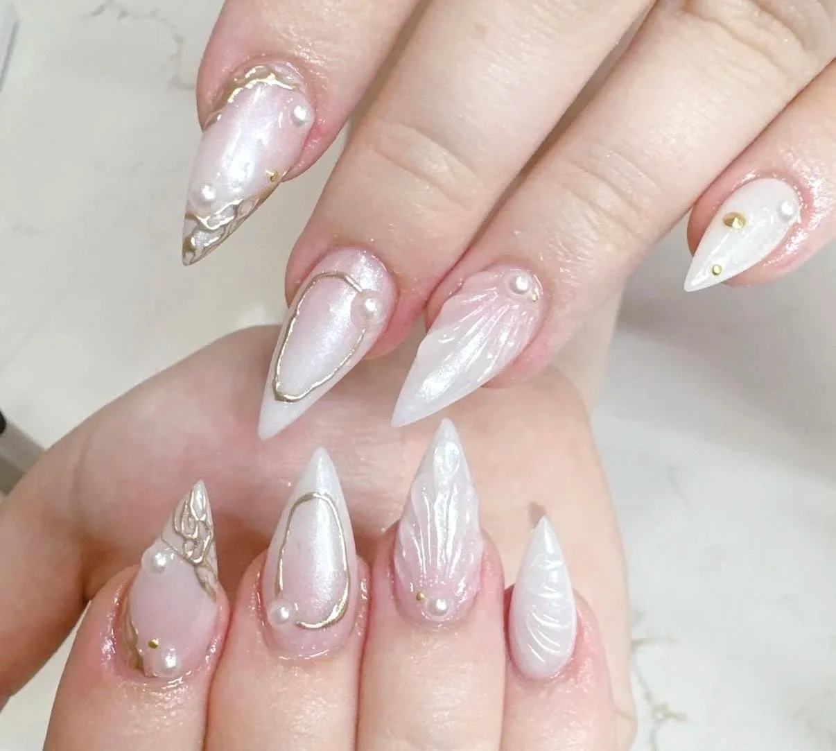 Close-up of decorated acrylic nails with white and pearlescent finishes, gold outlining, and pearl and gold embellishments.