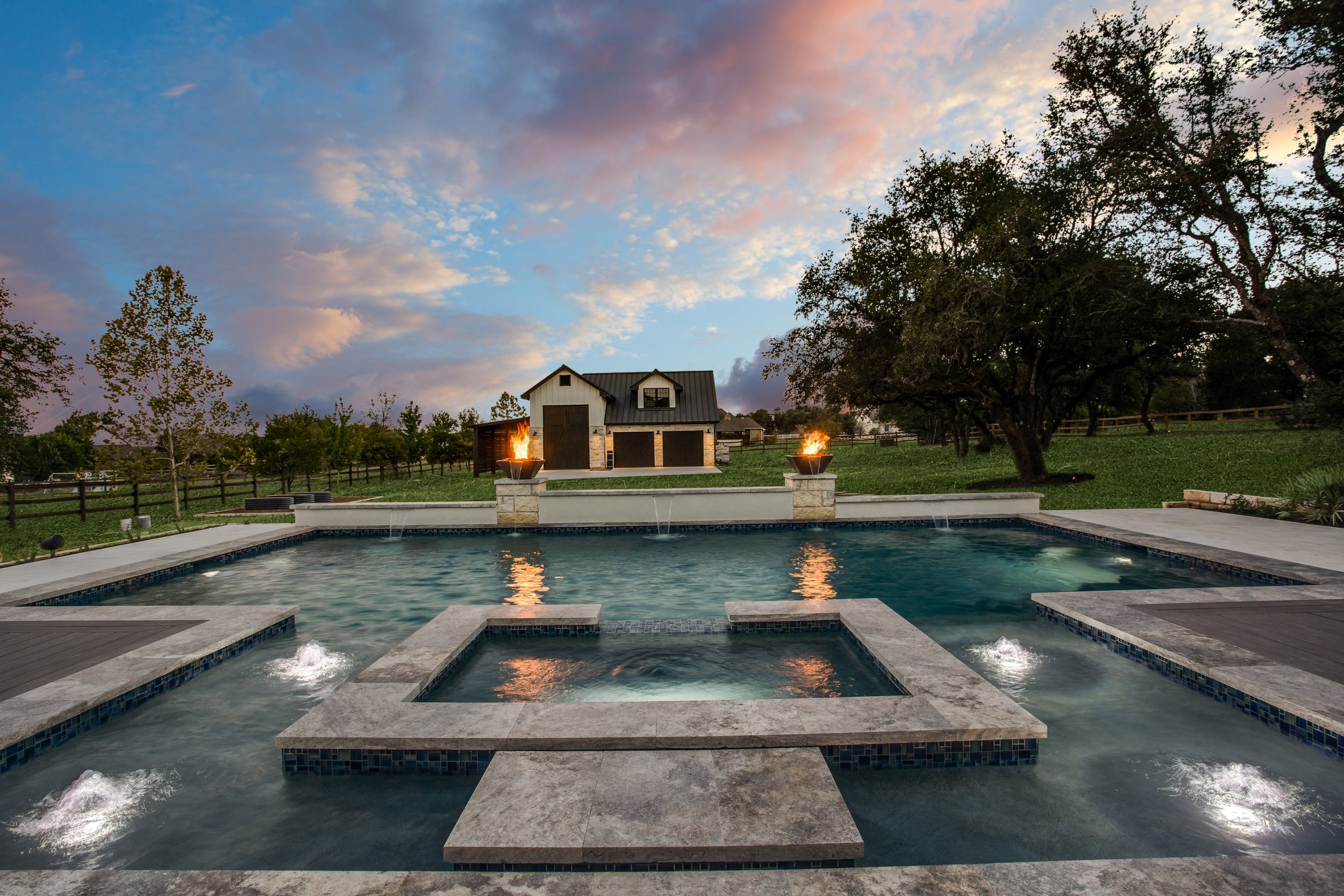 A backyard with a swimming pool and hot tub, a lawn, trees, and a barn-like building with fire features at sunset.