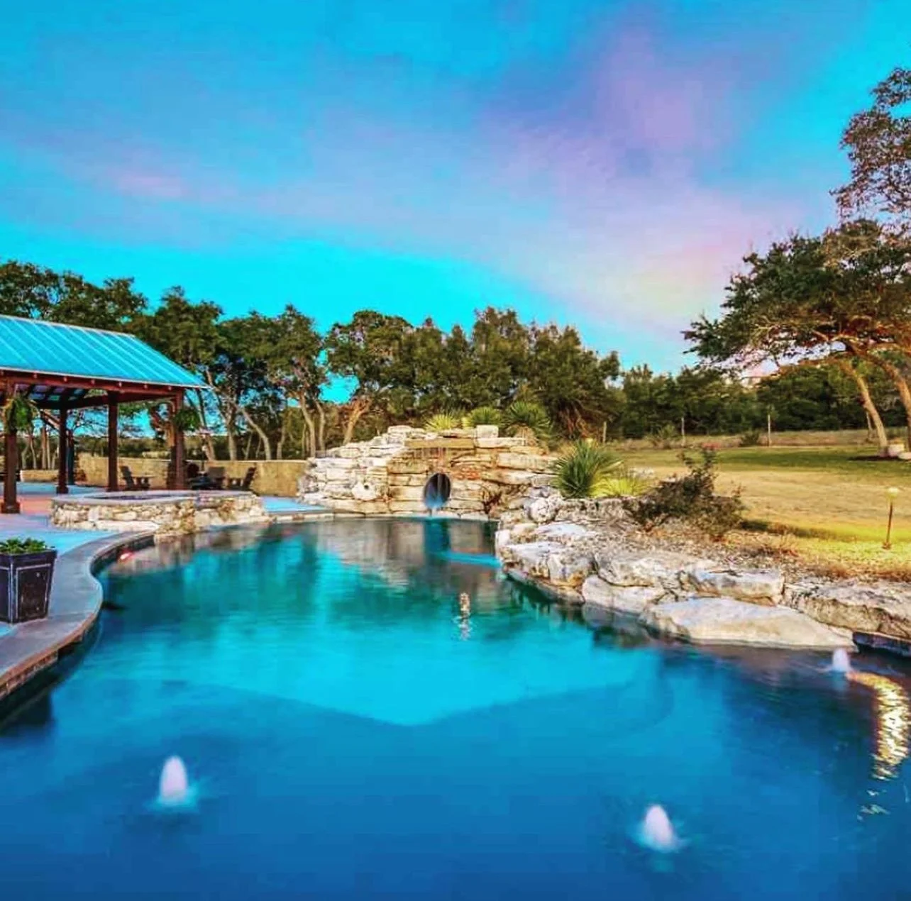 A serene outdoor swimming pool with a small waterfall feature, surrounded by trees and a clear blue sky.