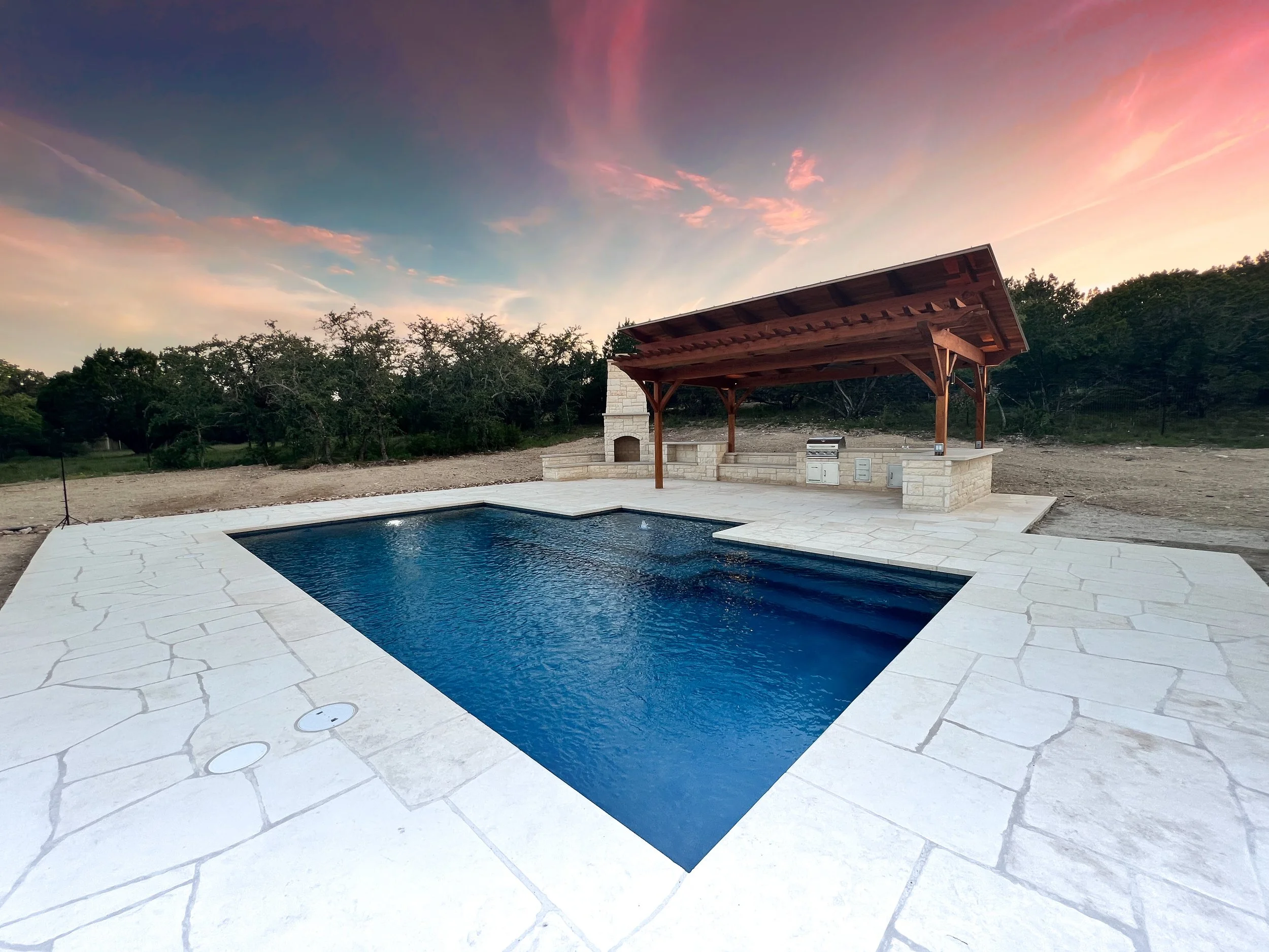 Empty backyard swimming pool with stone deck, wooden shelter with outdoor kitchen, and a sunset sky in the background.