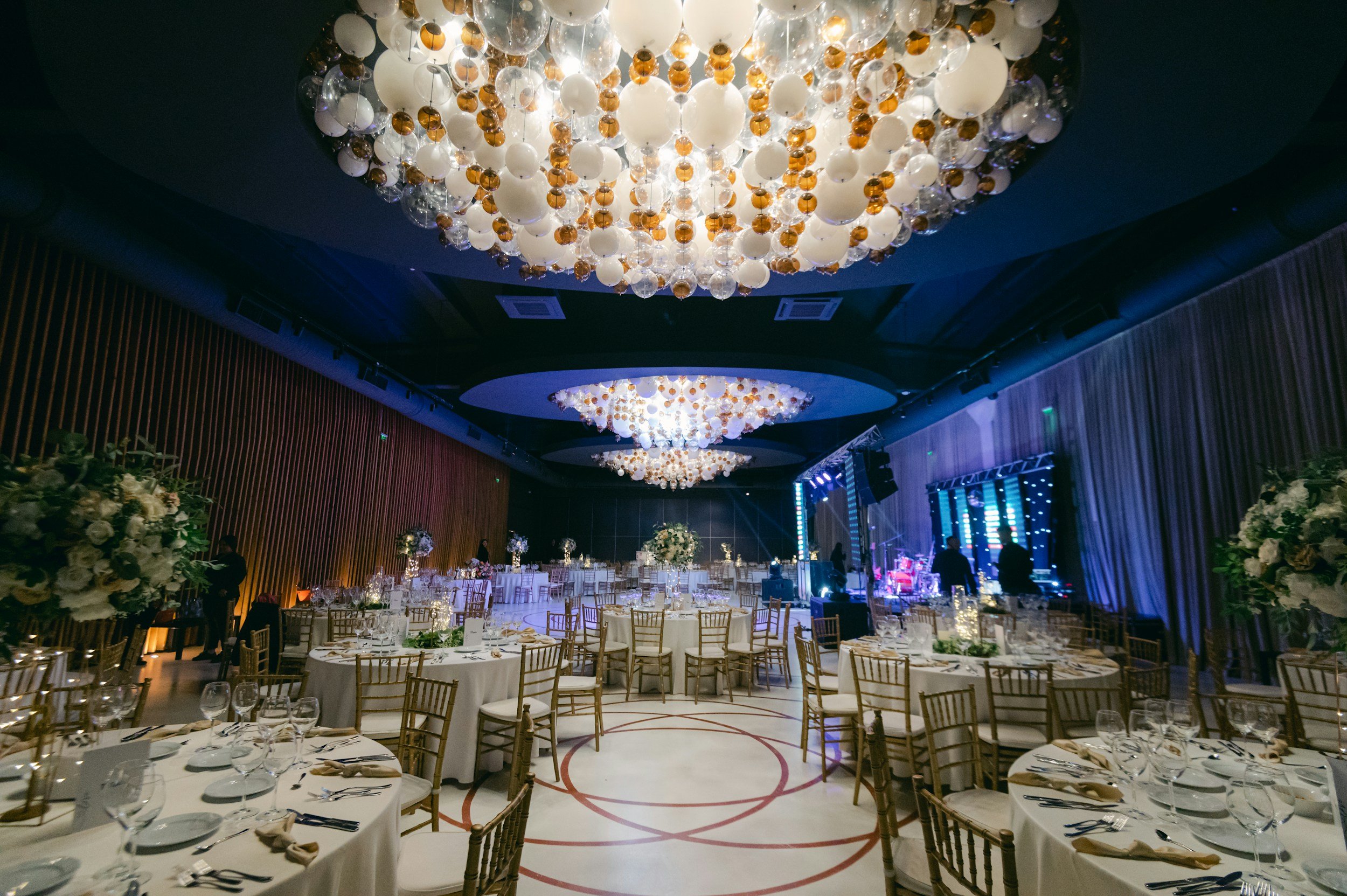 Elegant banquet hall decorated for a formal event with chandeliers hanging from the ceiling, round tables adorned with white tablecloths, floral centerpieces, and gold chairs, a stage with musical instruments and lighting setup.