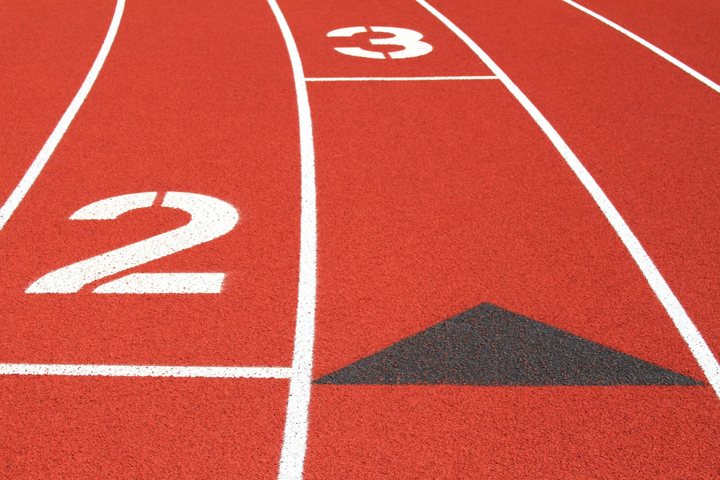A red running track with white lane markings, numbered 2 and 3, and a black triangular starting line marker in lane 2.