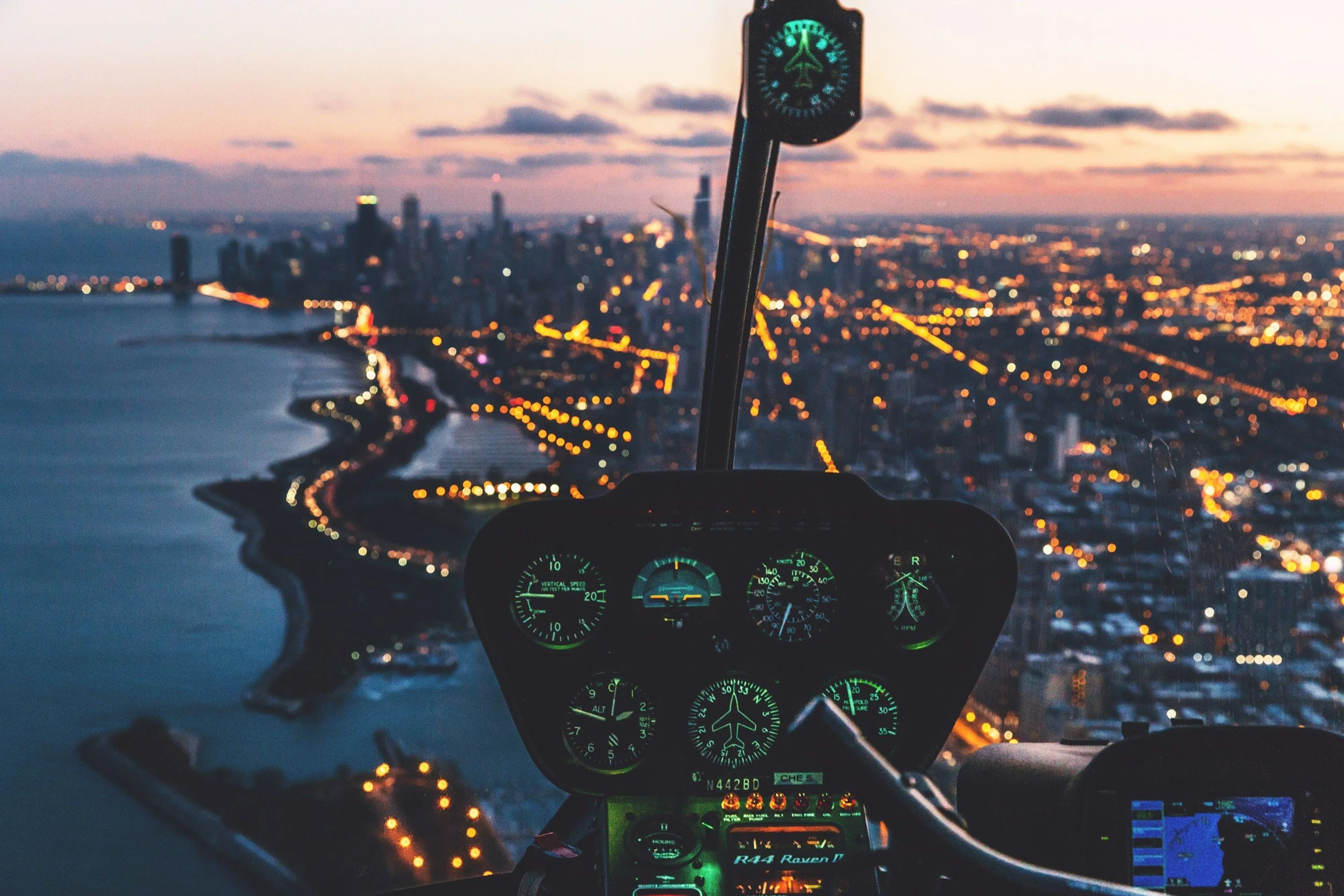 Cockpit view of a helicopter flying over a city at sunset, showing illuminated buildings and winding waterfront.