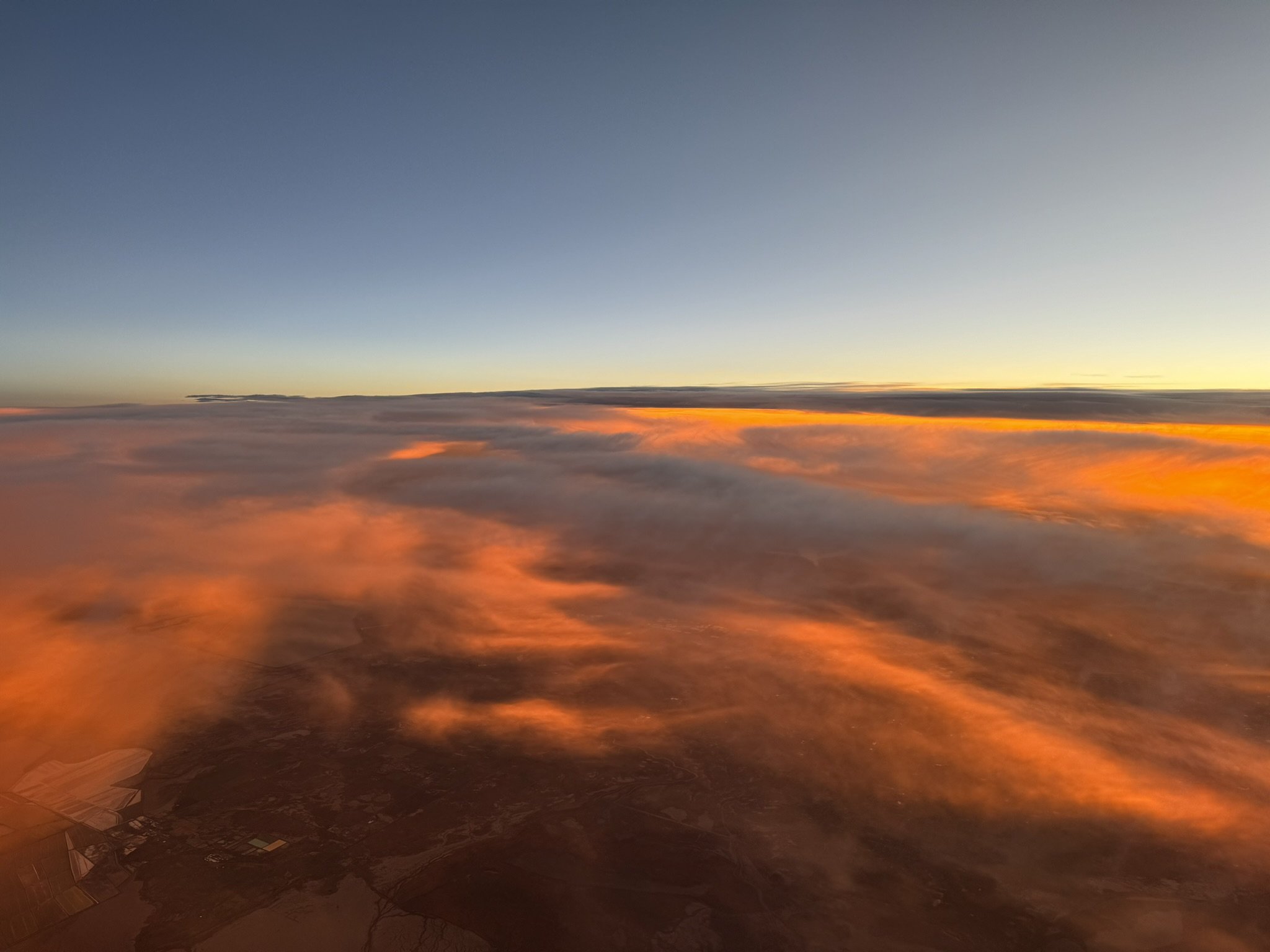 View from an airplane window showing a sunset sky with clouds illuminated in orange at high altitude.