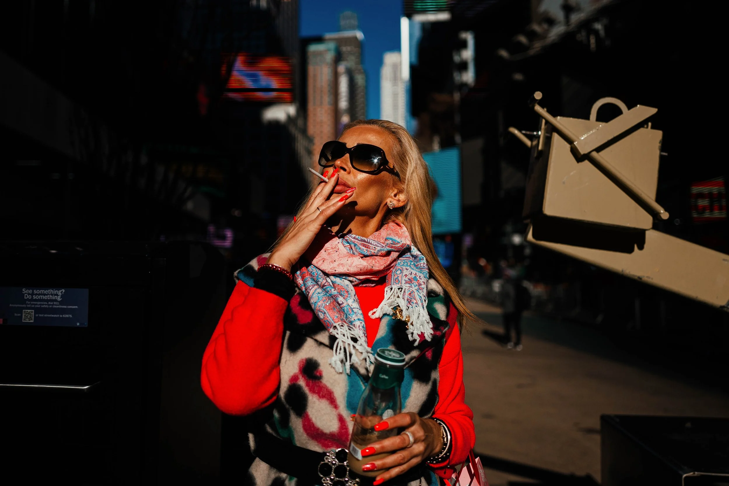 Mujer con gafas grandes y cabello rubio, sosteniendo una botella y fumando en una calle urbana, con edificios altos y pantallas digitales en el fondo.