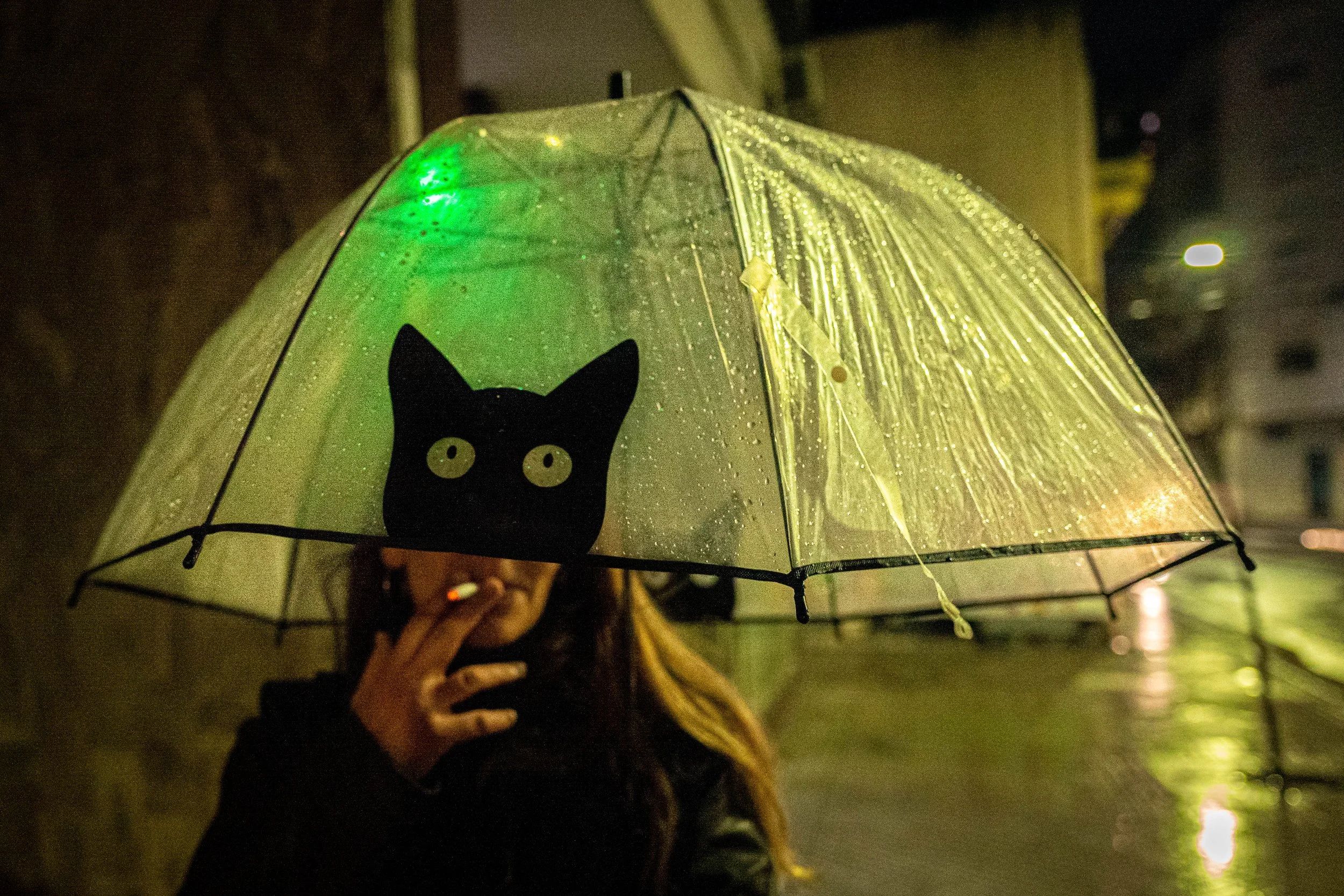 Persona sosteniendo un paraguas transparente con un dibujo de un gato negro y ojos amarillos, en un entorno lluvioso y nocturno.