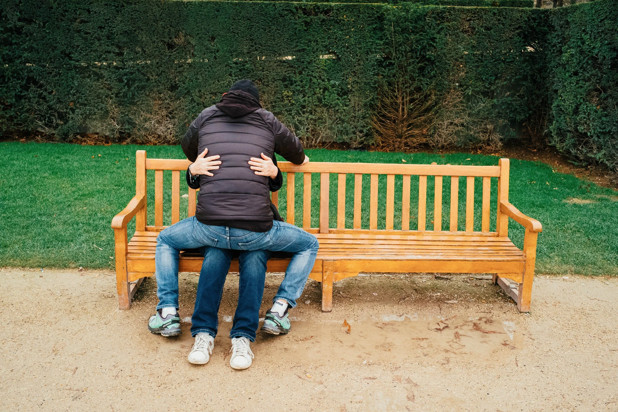 Dos personas en un parque, una abrazando a la otra mientras están sentadas en un banco de madera, con fondo de arbustos verdes.
