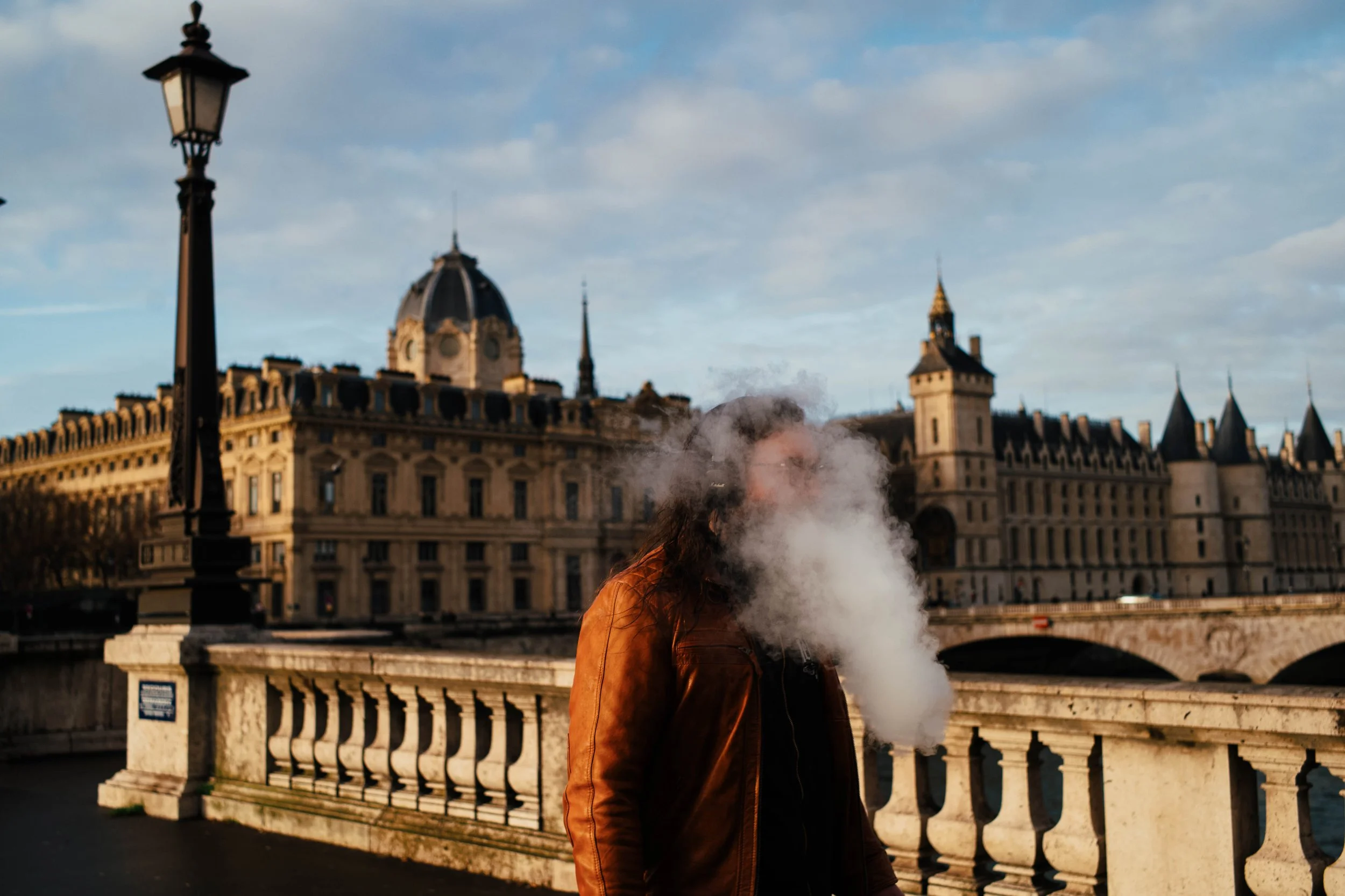 Persona expulsando vapor frente a un castillo en París, con puente y lámpara en primer plano.