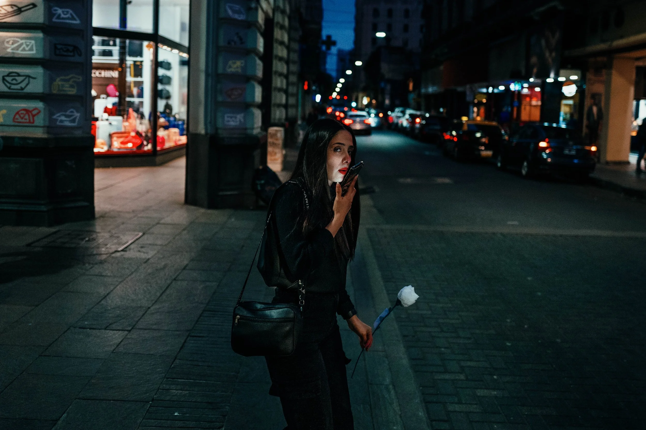 Mujer con mochila negro, sosteniendo una rosa y usando un teléfono móvil en una calle urbana por la noche