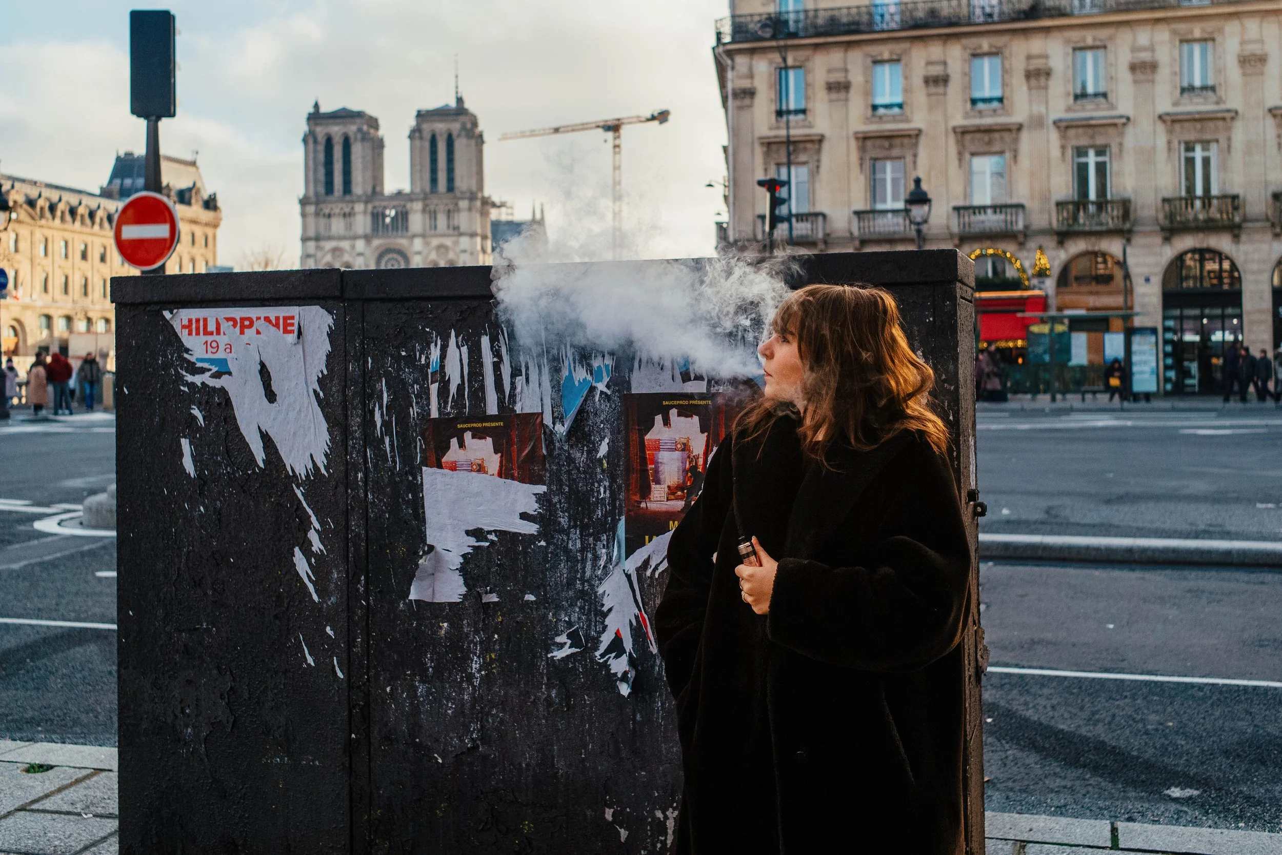 Mujer con abrigo negro fumando en la calle con edificios históricos y tráfico en el fondo
