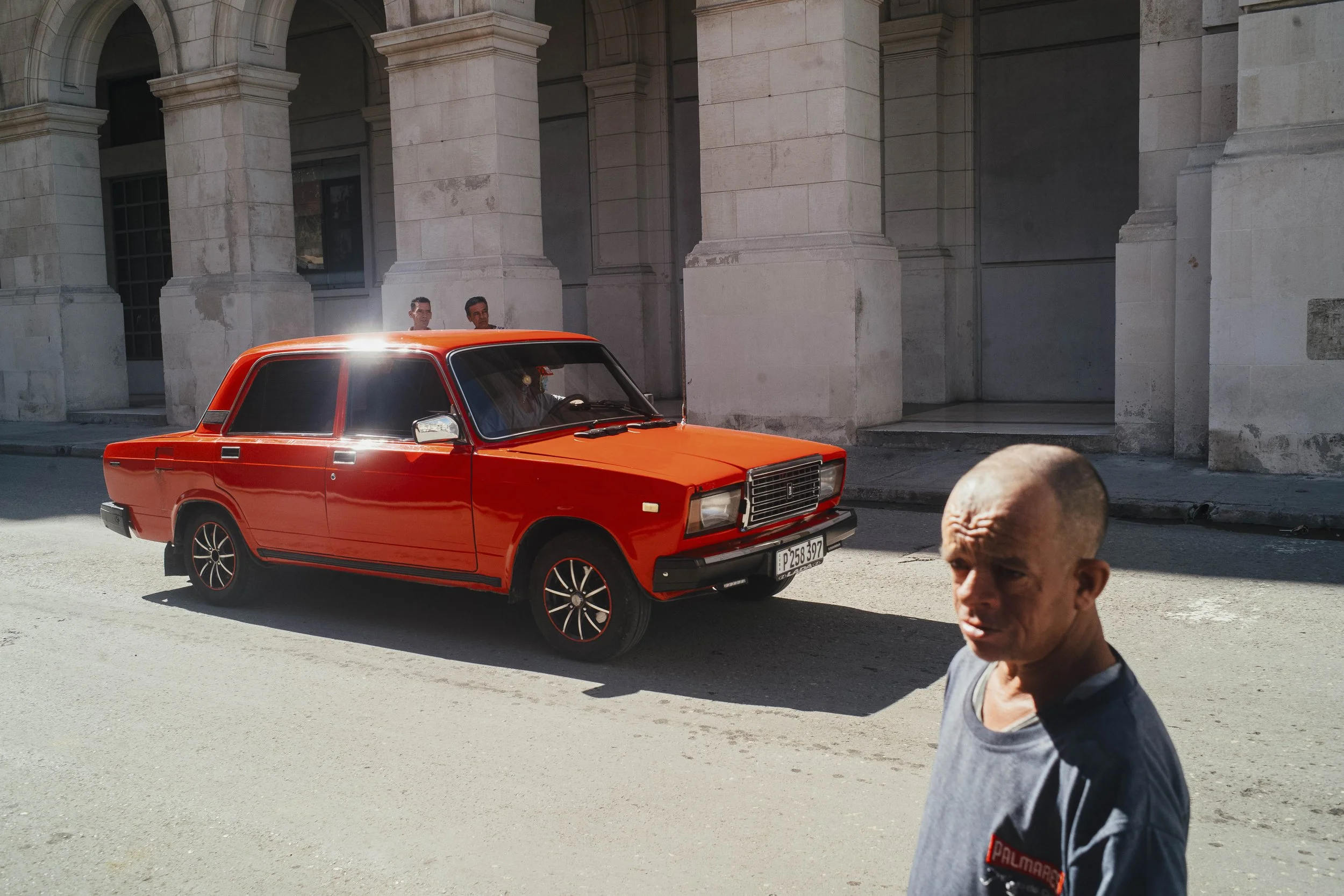 Un hombre calvo con camiseta gris mira hacia la cámara en una calle en la ciudad, con un coche rojo estacionado y un edificio de estilo clásico en el fondo.