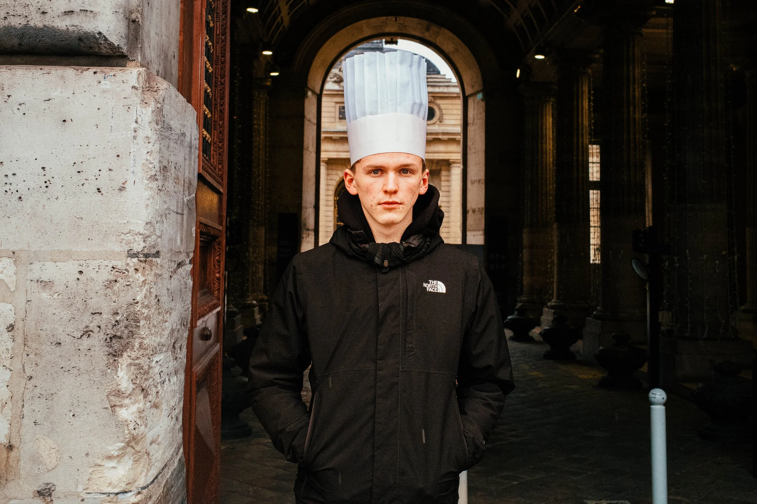Un joven con chaqueta negra y gorro de chef posando en una calle con arcos y edificios históricos.
