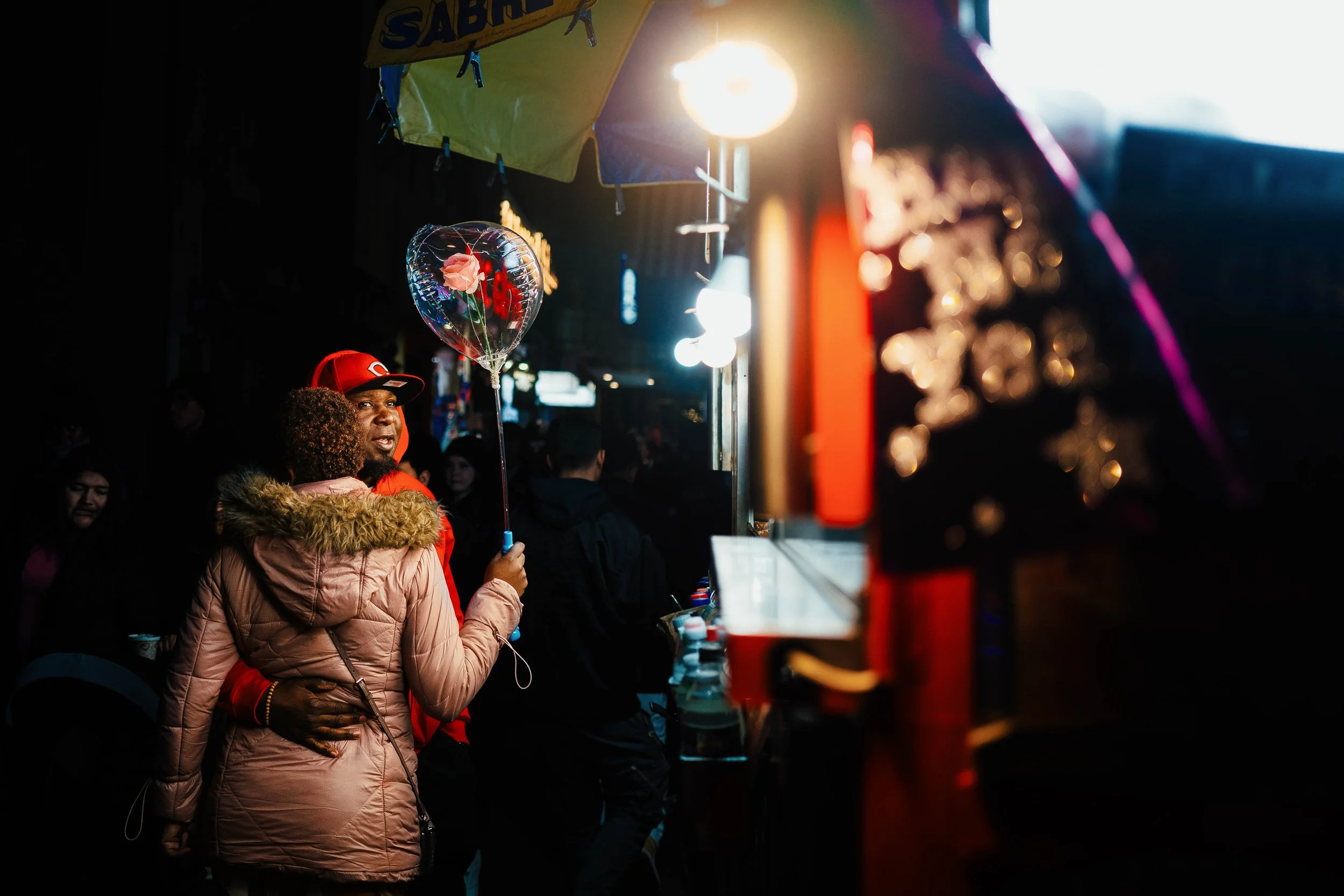 Una pareja abrazada en una feria nocturna, la mujer sostiene un globo en forma de corazón con una rosa en su interior.