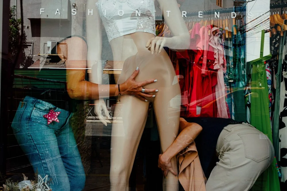 Ventana de tienda de ropa con maniquí sin ropa y dos personas organizando prendas. Visible ropa y accesorios en exhibición.