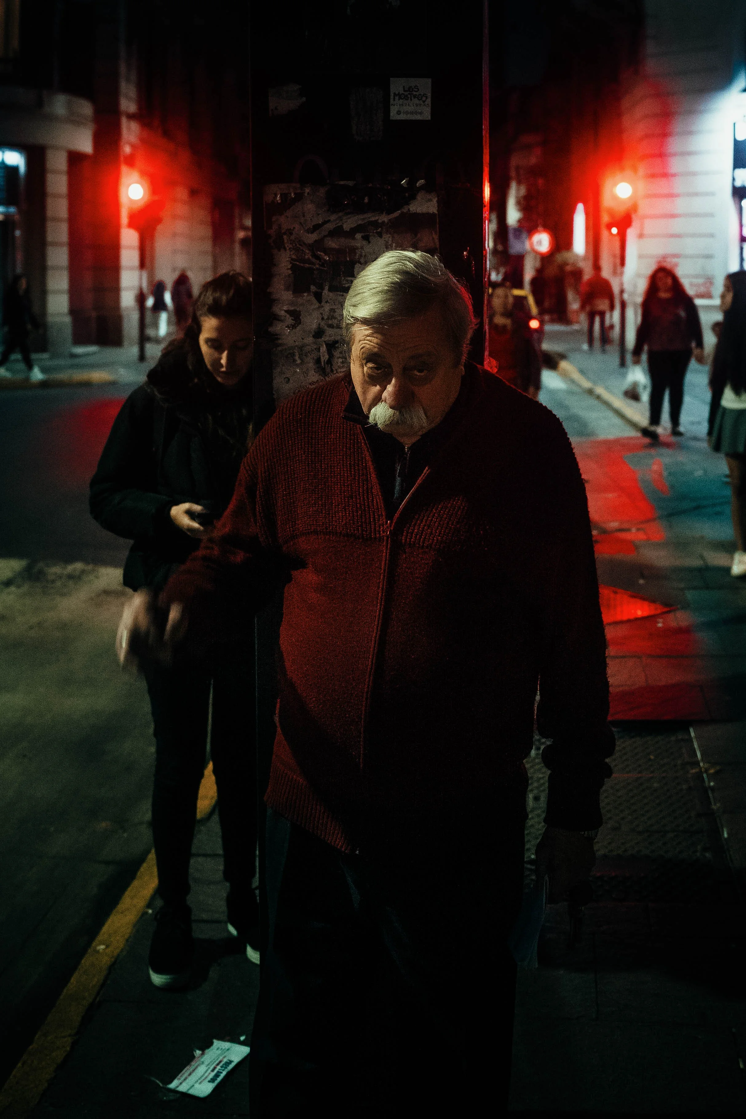 Hombre mayor con barba y pelo canoso, usando un suéter rojo oscuro, caminando en la calle por la noche, con luces rojas y personas en el fondo.