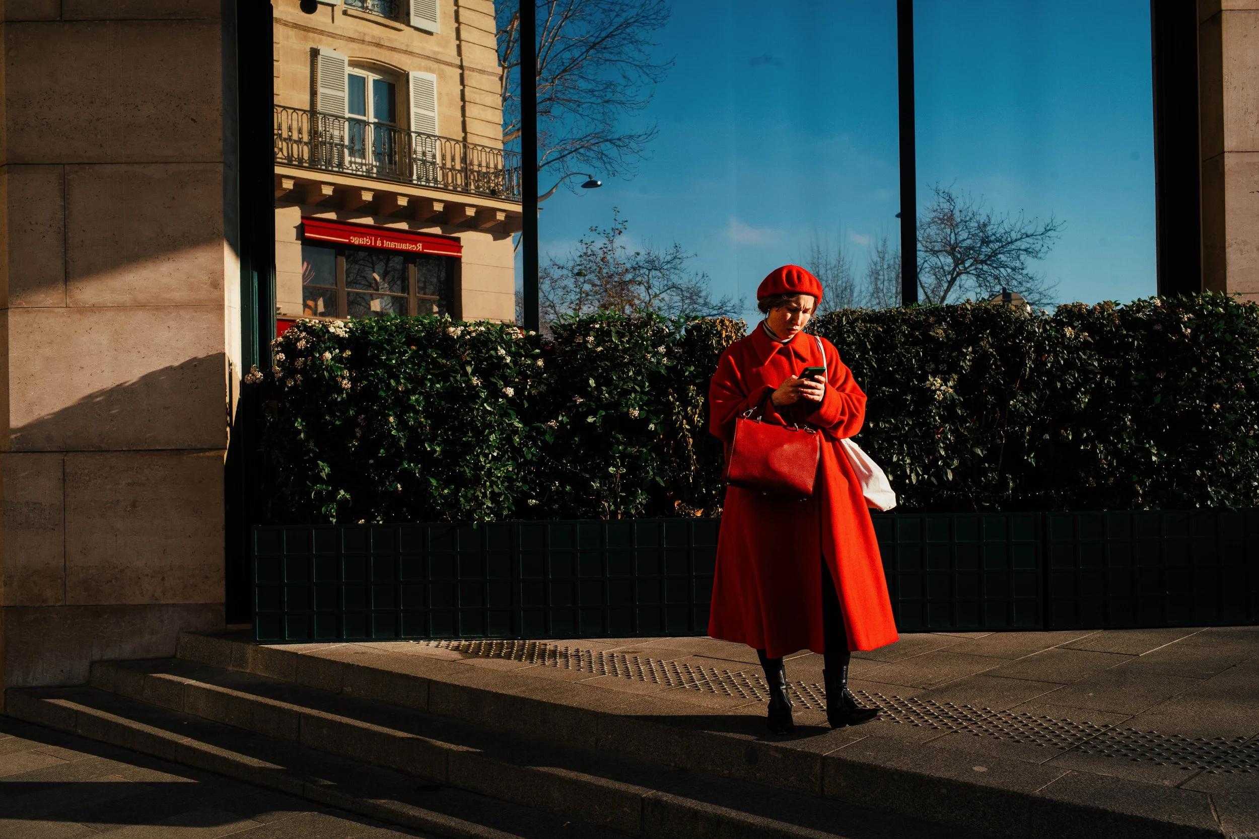 Una mujer vestida completamente de rojo, con un sombrero, abrigo largo y bolso, mirando su teléfono en una ciudad bajo el sol, con árboles sin hojas y un edificio de fondo.