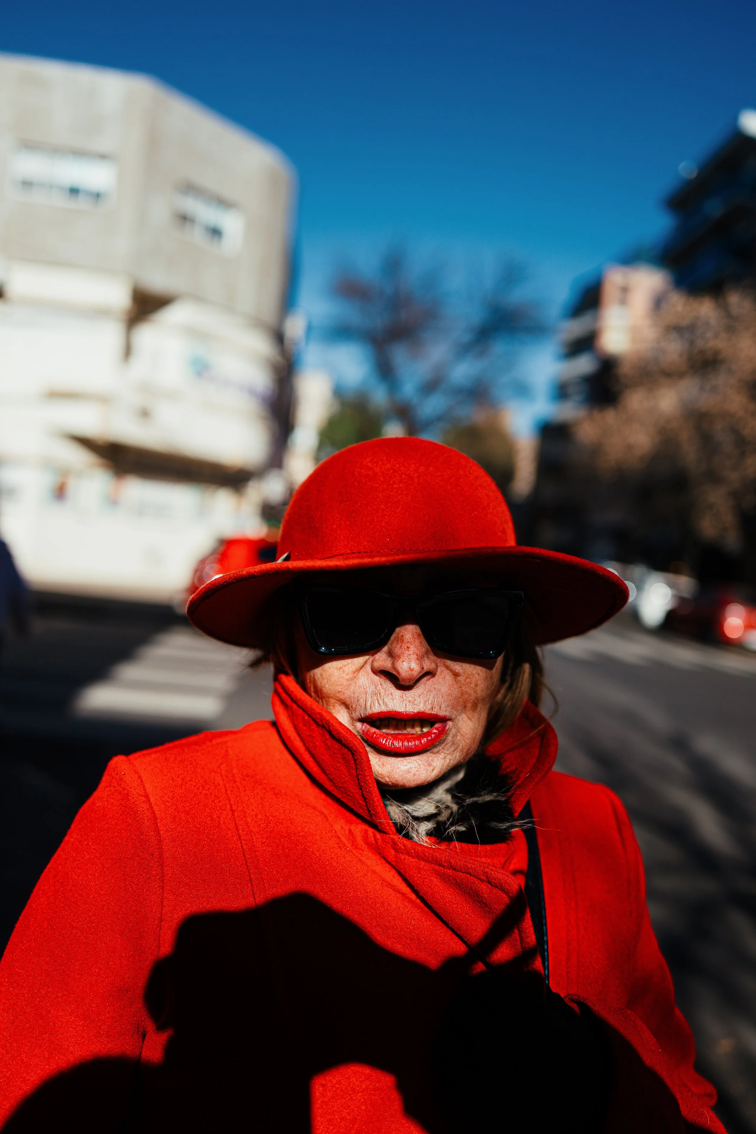Mujer con sombrero y abrigo rojos en la calle con edificios y árboles al fondo, vestida con gafas oscuras y maquillaje visible, en un día soleado.