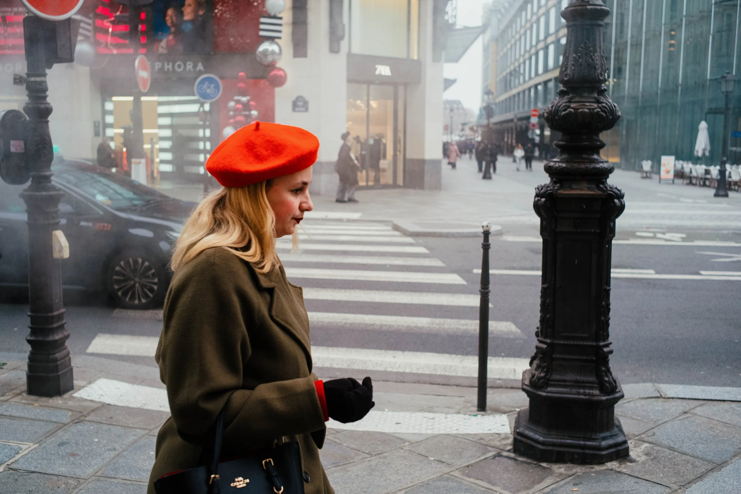 Mujer con sombrero rojo y abrigo verde caminando por una acera en una ciudad con calles transitadas y tiendas.