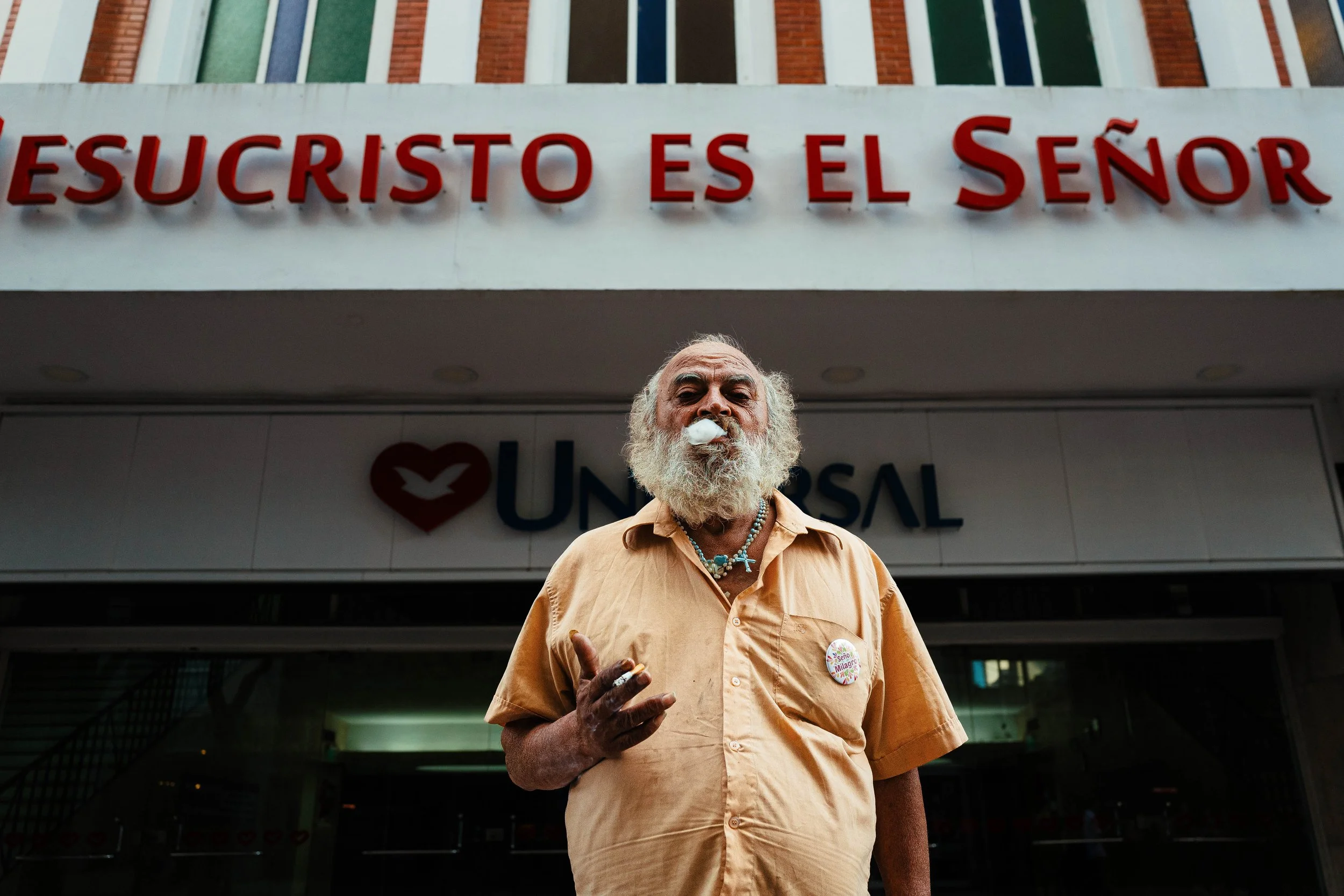 Un hombre de barba blanca y cabello a rayas, usando una camiseta de color amarillo claro, de pie frente a un edificio con letreros en español. Tiene una etiqueta en el pecho y una llamada en la mano, mientras mira a la cámara. El letrero en la parte 