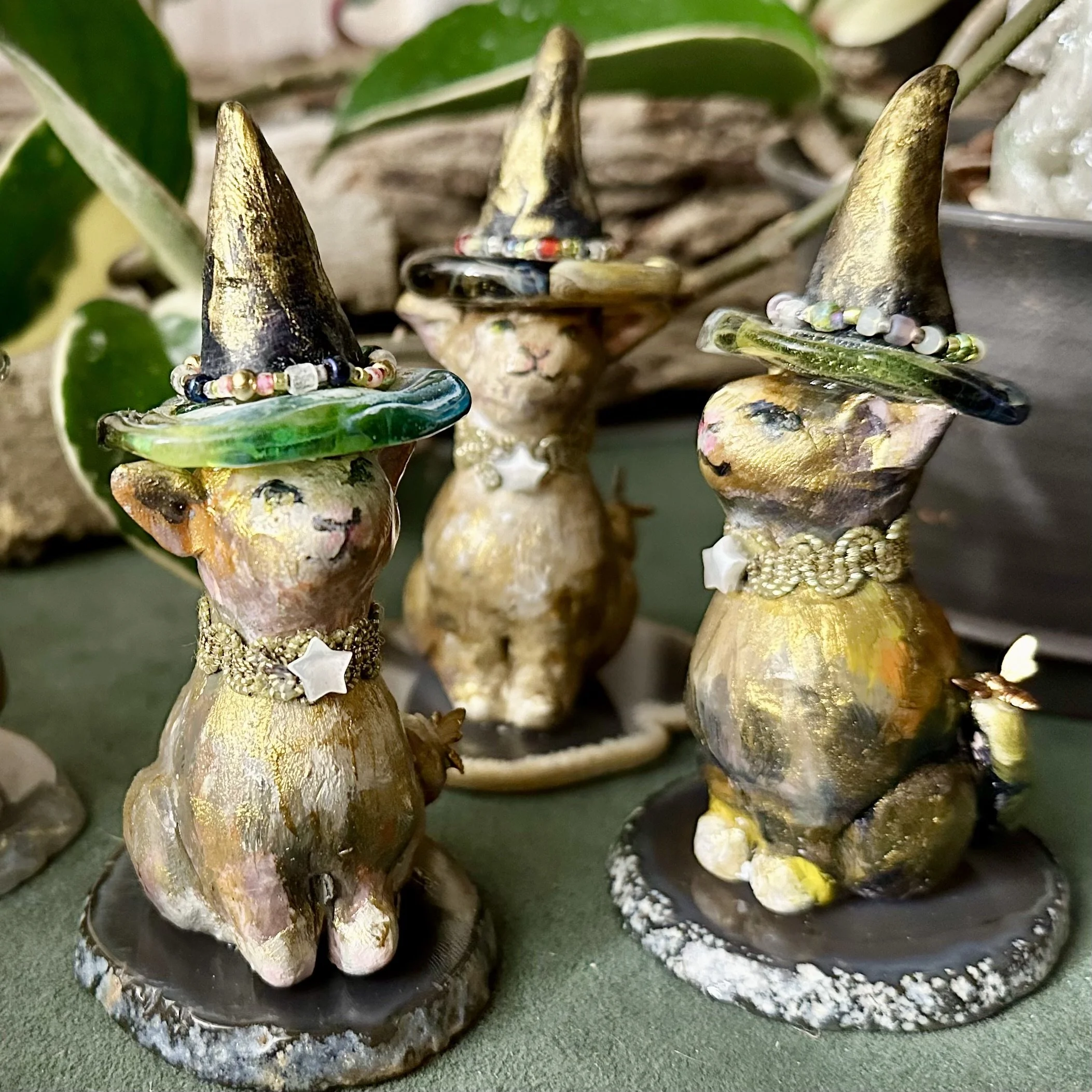 Three whimsical rabbit figurines dressed as witches with pointed hats, necklaces, and painted faces, placed on a green surface with plants in the background.