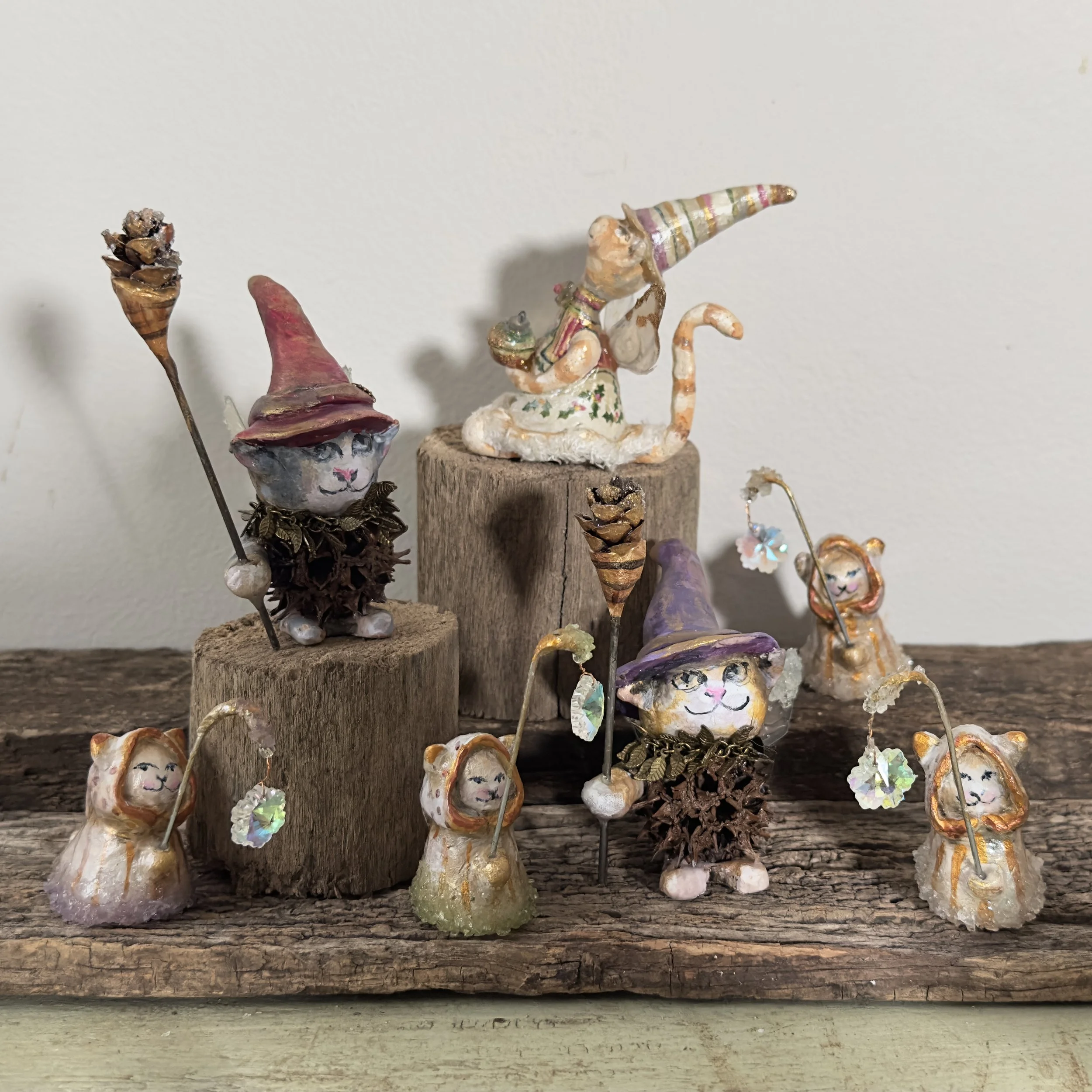 Collection of small, whimsical figurines of cats and mice dressed as witches, with some holding flower or star-shaped wands, arranged on wooden blocks and a rustic wood surface.