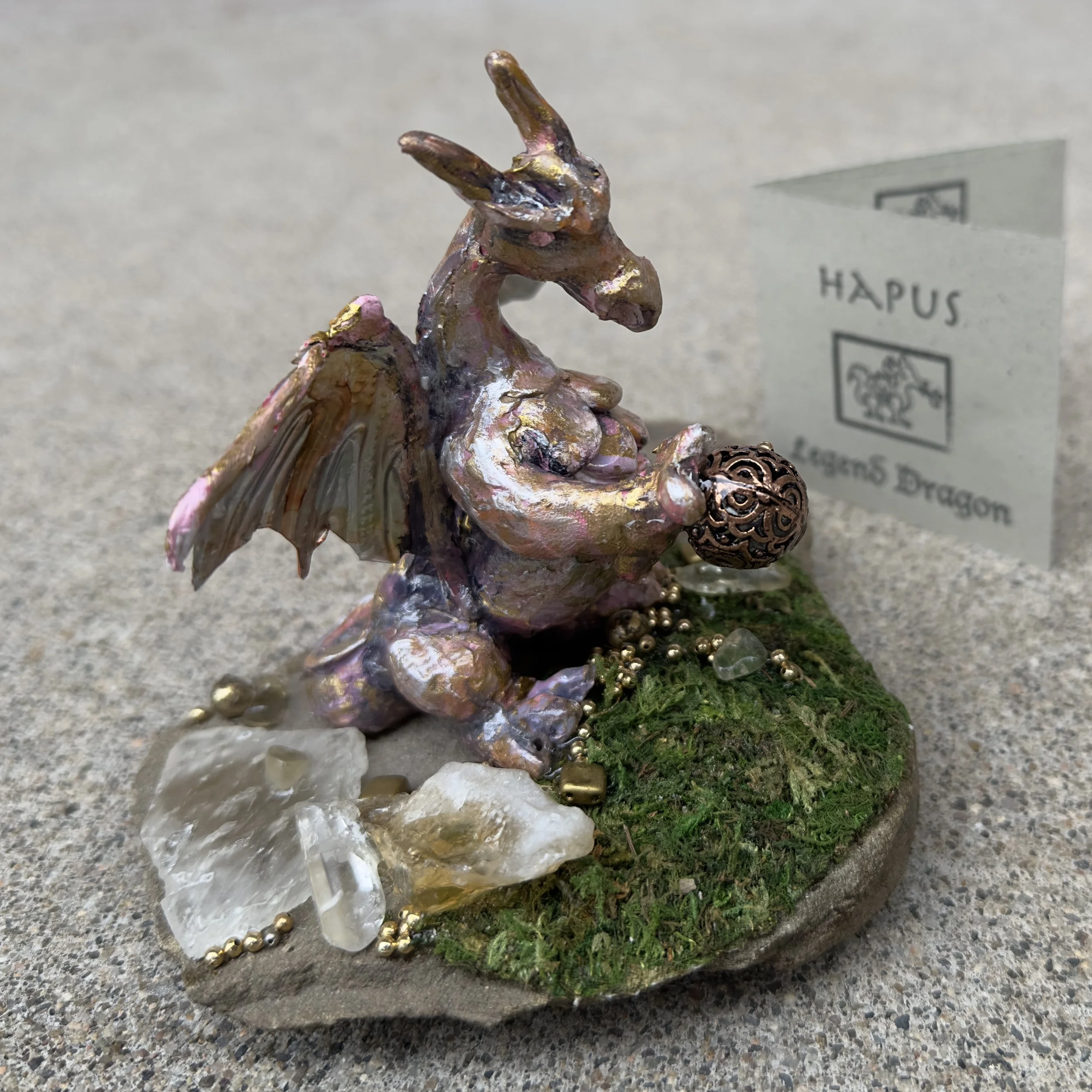 Fantasy sculpture of a dragon holding a decorative orb, with moss, quartz, and gold beads on a circular base, and a sign that reads 'HAPUS' and 'Eagon5 Dragon' in the background.