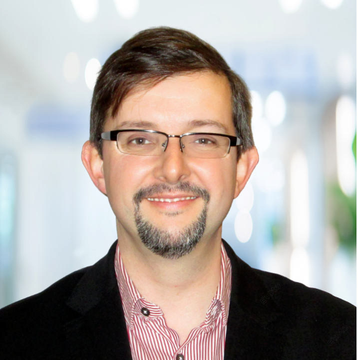 A Professional headshot middle-aged of Colm White Chief Program Officer at Caldera Therapeutics with short brown hair and a beard, wearing glasses, a black blazer, and a red striped dress shirt, smiling in an indoor setting with bright lighting.