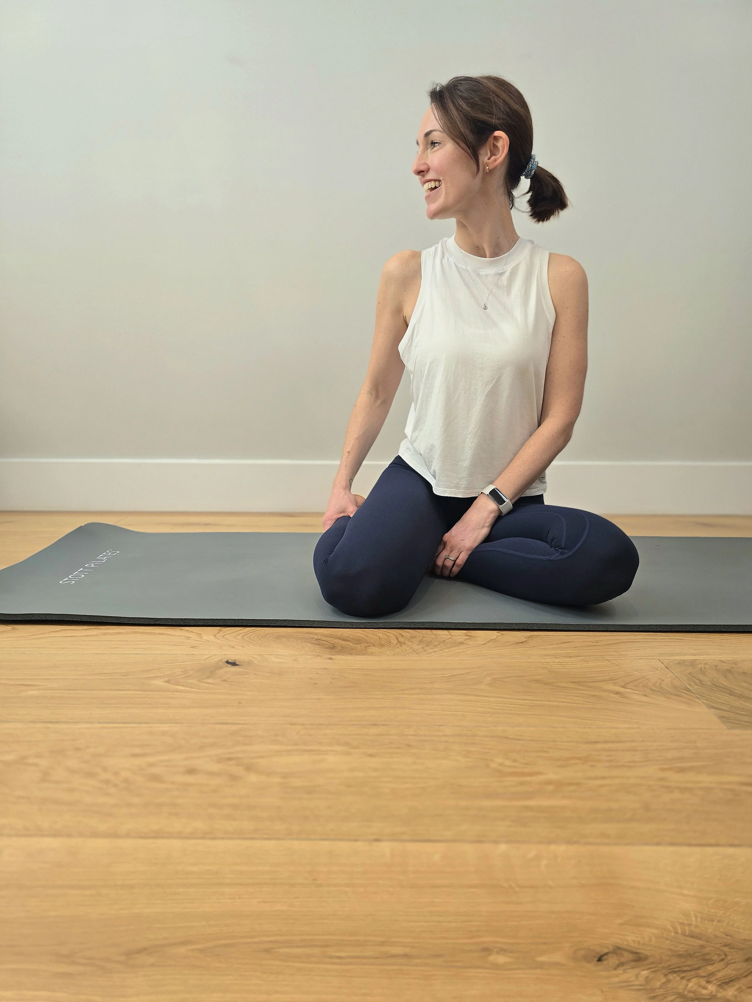 A woman with brown hair tied in a ponytail, wearing a white sleeveless top and navy leggings, is sitting on a yoga mat on a wooden floor, smiling and looking to her right in a yoga or stretching pose.