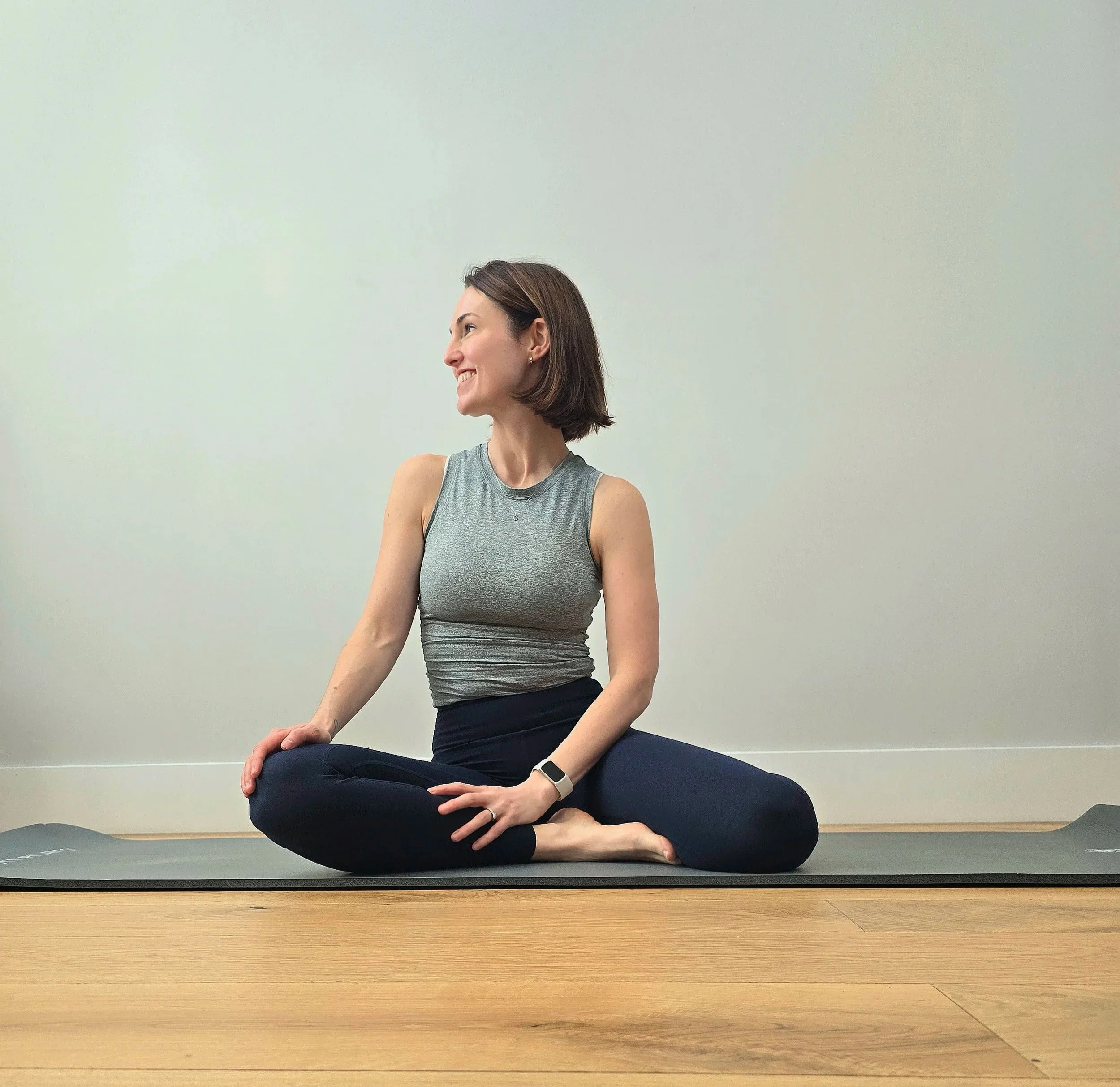 A woman sitting cross-legged on a yoga mat, smiling, with short brown hair, wearing a gray sleeveless top, black leggings, and a smartwatch, in a room with a light green wall and wooden floor.