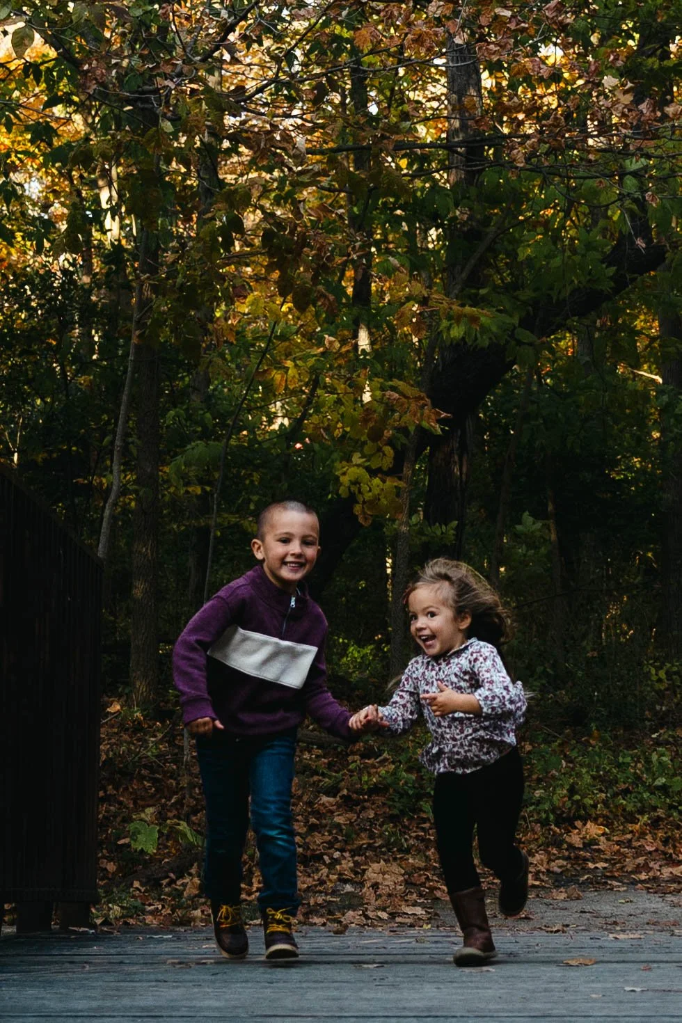 Two children, a boy and a girl, are playing and holding hands on a wooden bridge in a wooded area with fall foliage.
