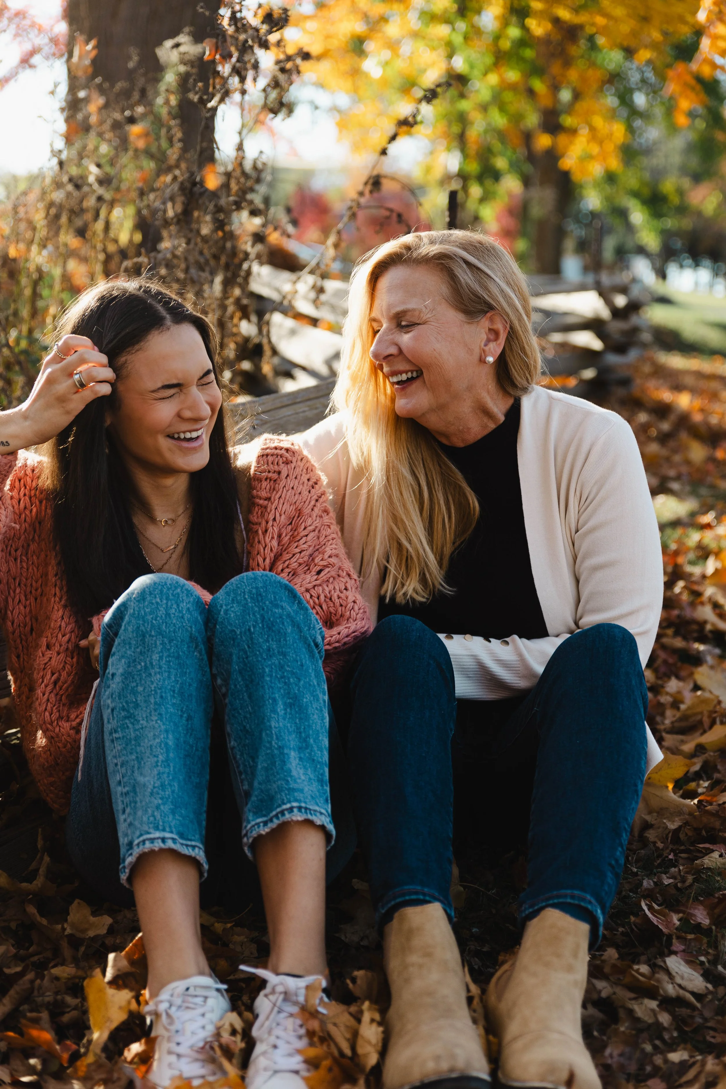 Two women sitting on the ground with autumn leaves, smiling and laughing outdoors.
