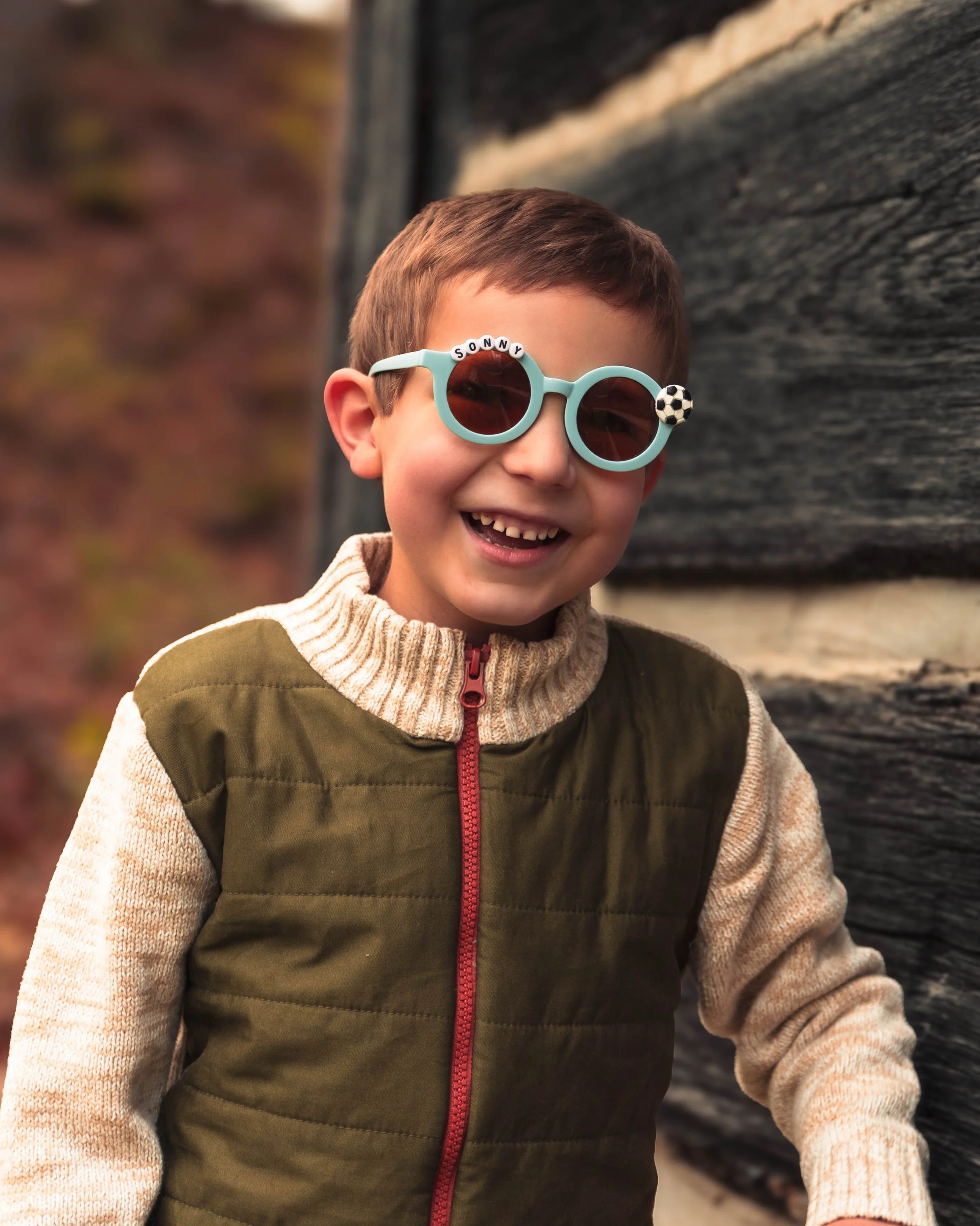 A young boy with brown hair smiling, wearing sunglasses with the word 'Sunny' and a small soccer ball on the sides, dressed in a green and beige hoodie, standing outdoors near a textured dark wooden wall.