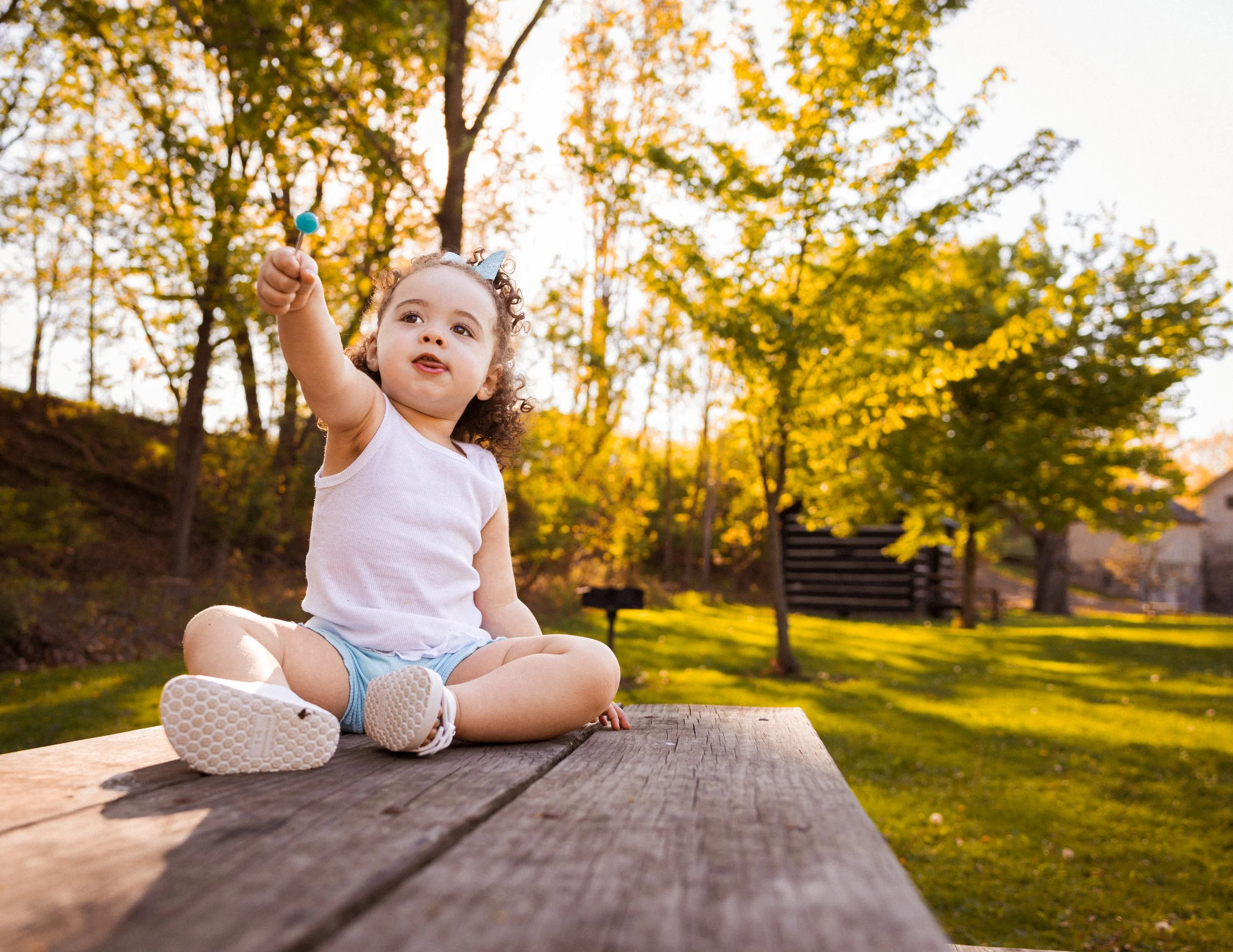 A young girl sitting cross-legged outdoors on a wooden surface during fall, holding a blue lollipop, with trees and autumn foliage in the background.