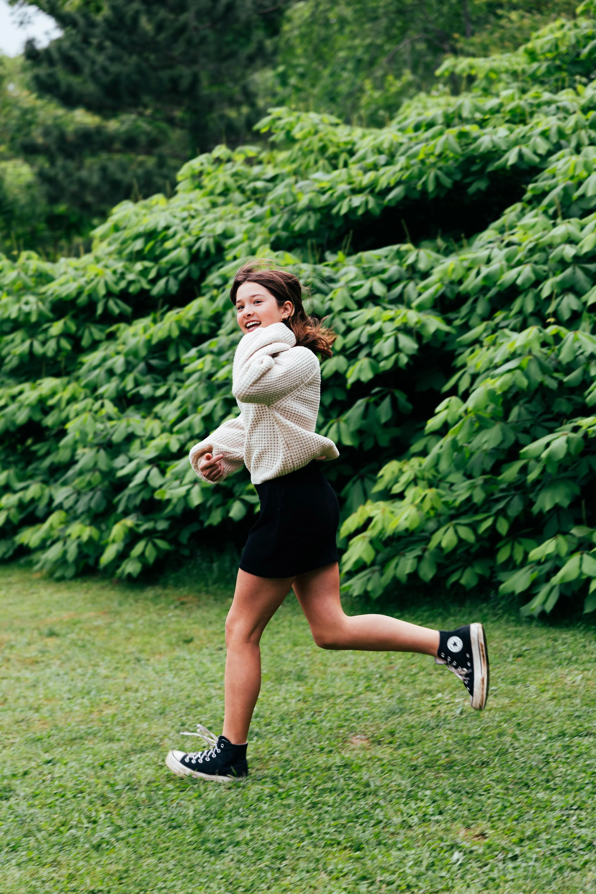 A girl running on a grassy field, wearing a beige sweater, black skirt, and black sneakers with white soles, with a large green leafy bush in the background.