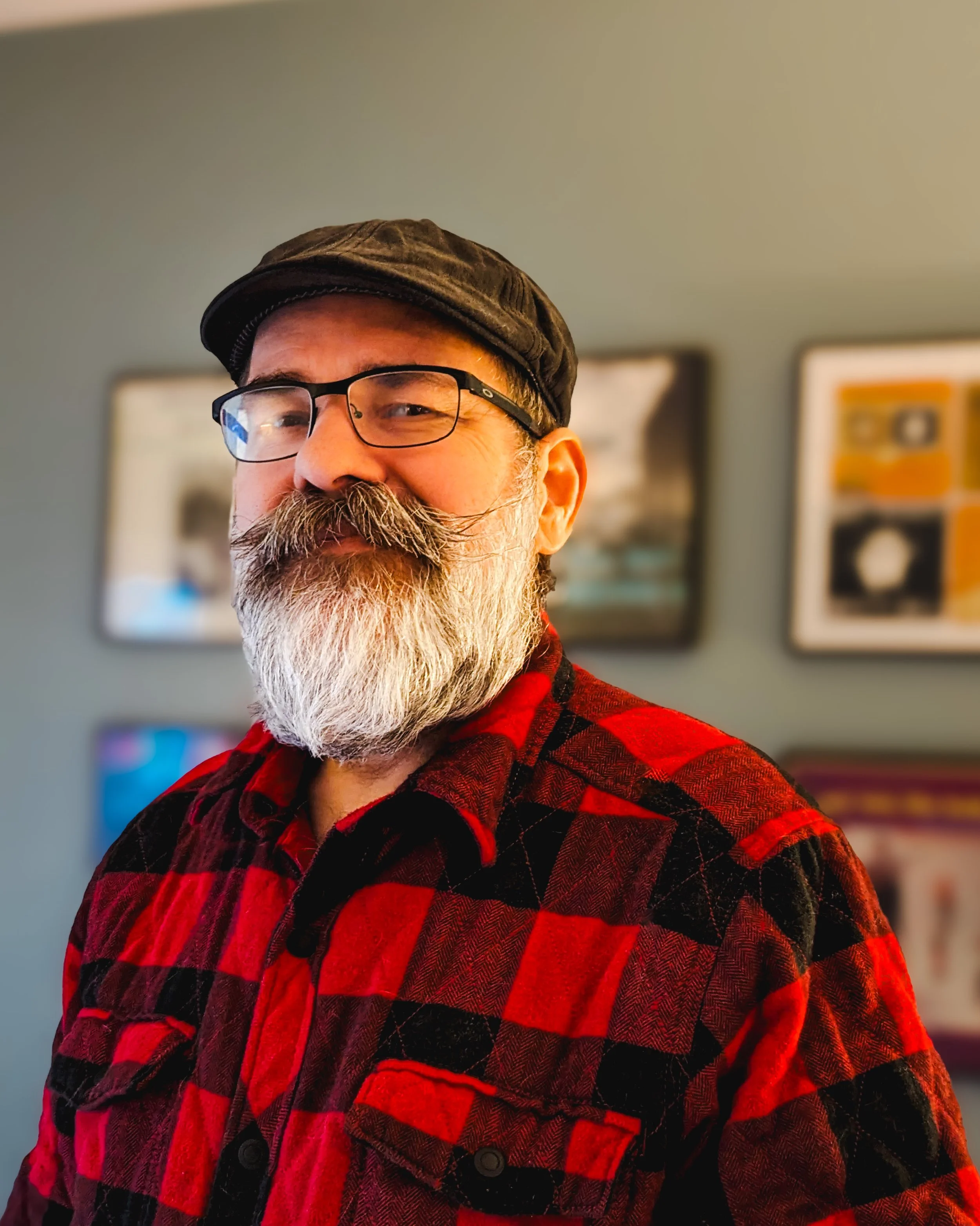 Close-up of a smiling man with glasses, a gray beard, a black cap, wearing a red and black plaid shirt, standing indoors with framed pictures on a light gray wall behind him.