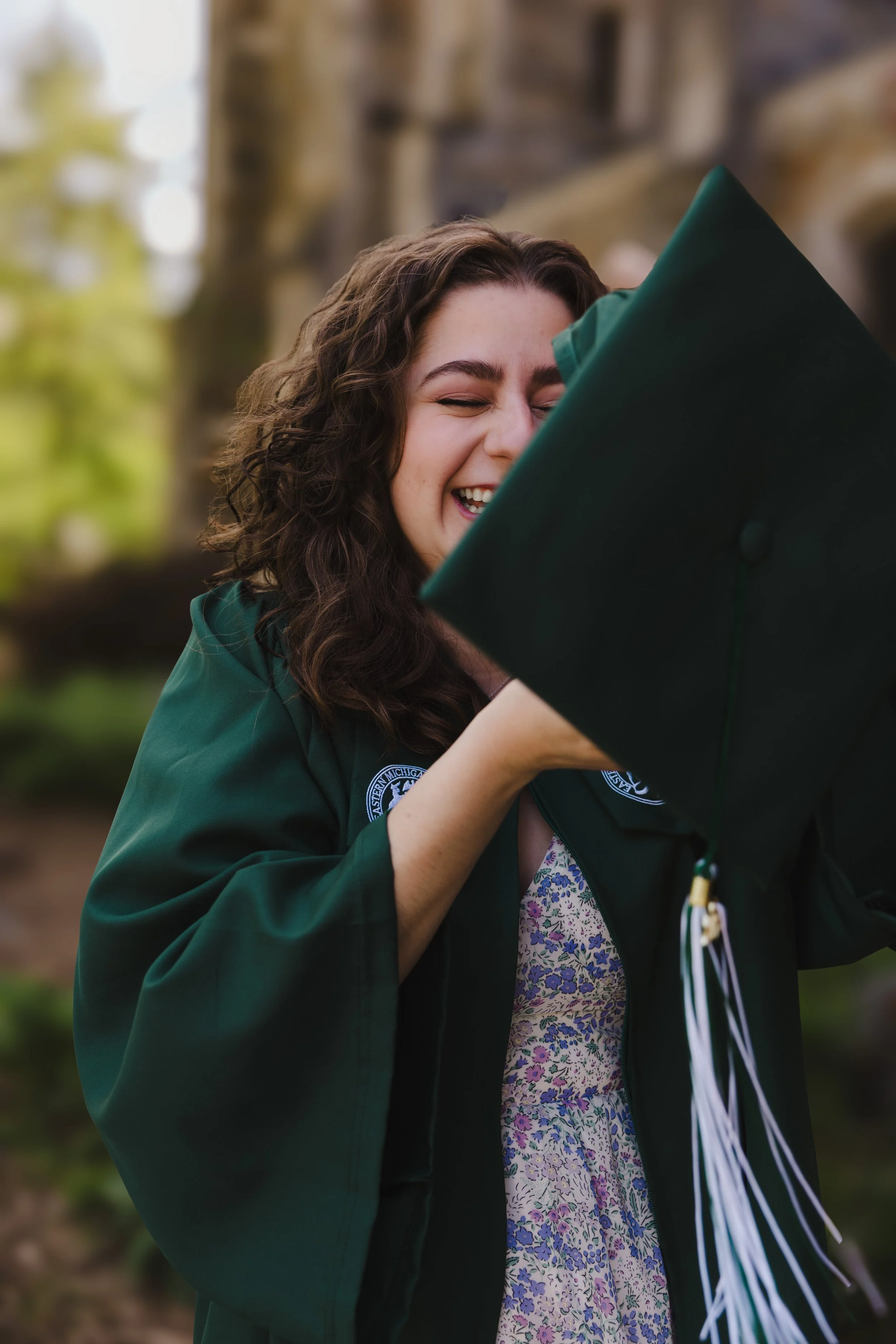 A young woman with curly brown hair smiling and wearing a green graduation gown and cap, holding a diploma, outdoors with blurred trees in the background.