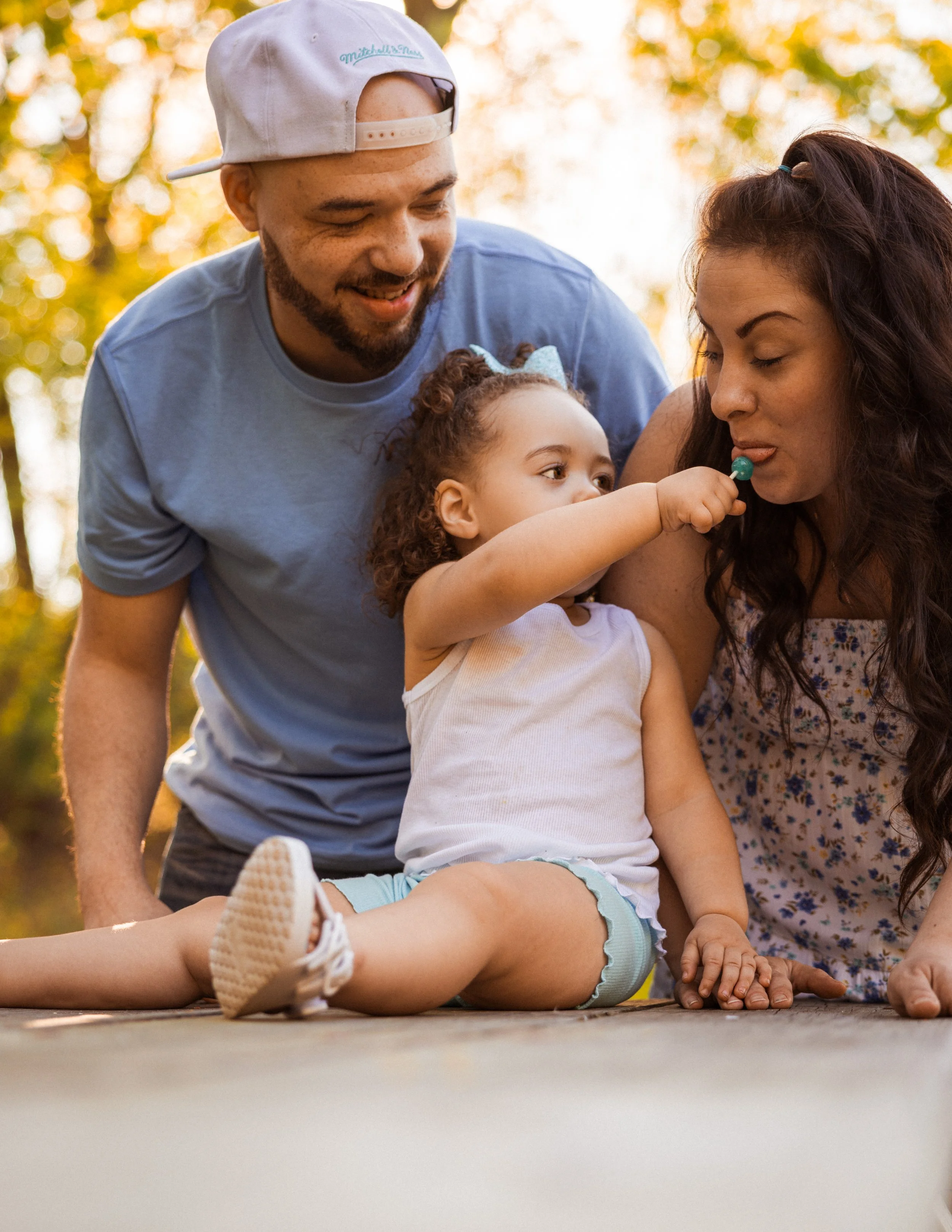 A family enjoying a day outdoors with a young girl lying on the ground, two adults leaning over her, one holding her arm, in a park with trees and sunlight.