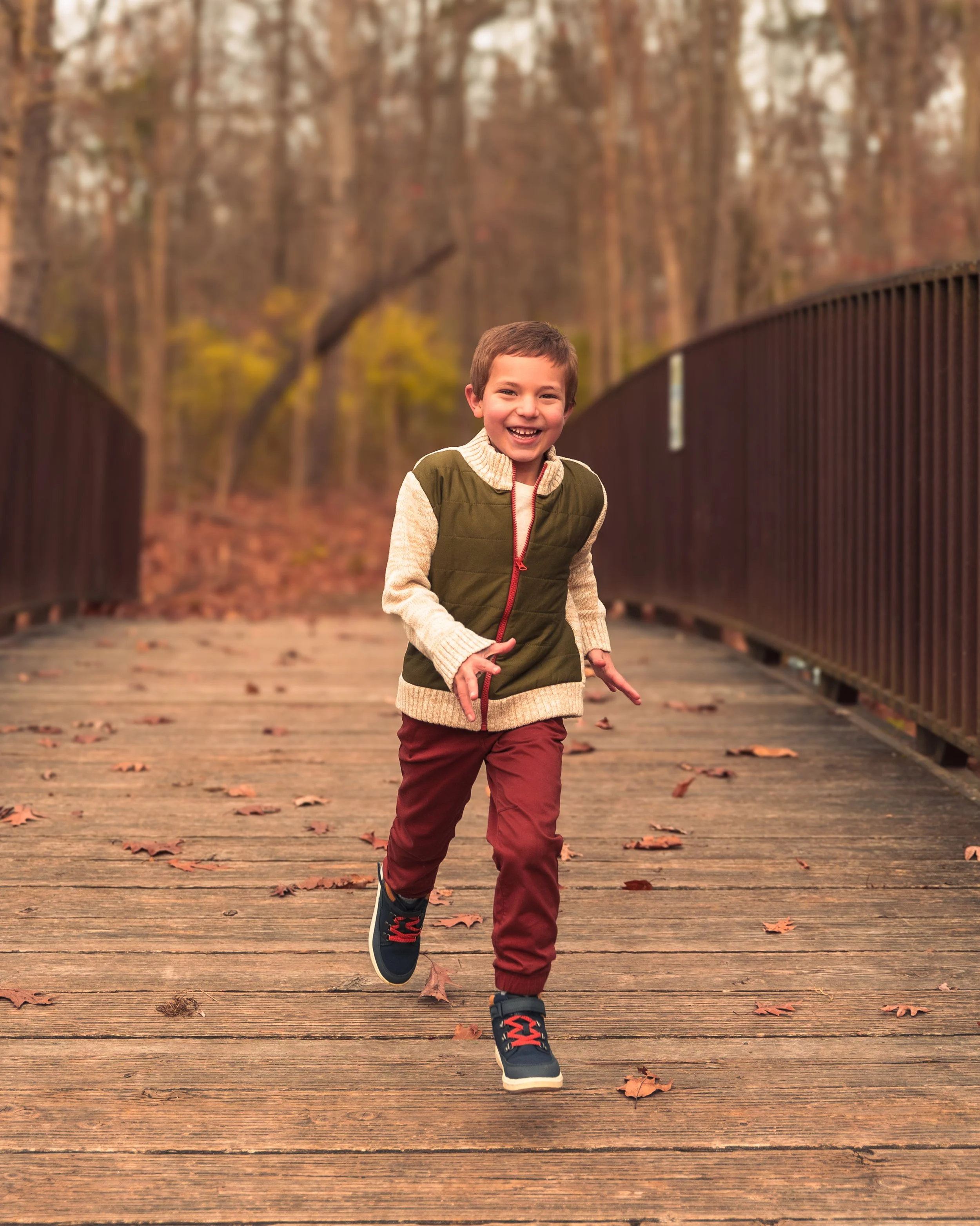 A young boy with short brown hair and a big smile running on a wooden bridge in a fall forest setting, wearing a green and beige jacket, maroon pants, and black shoes with red laces.